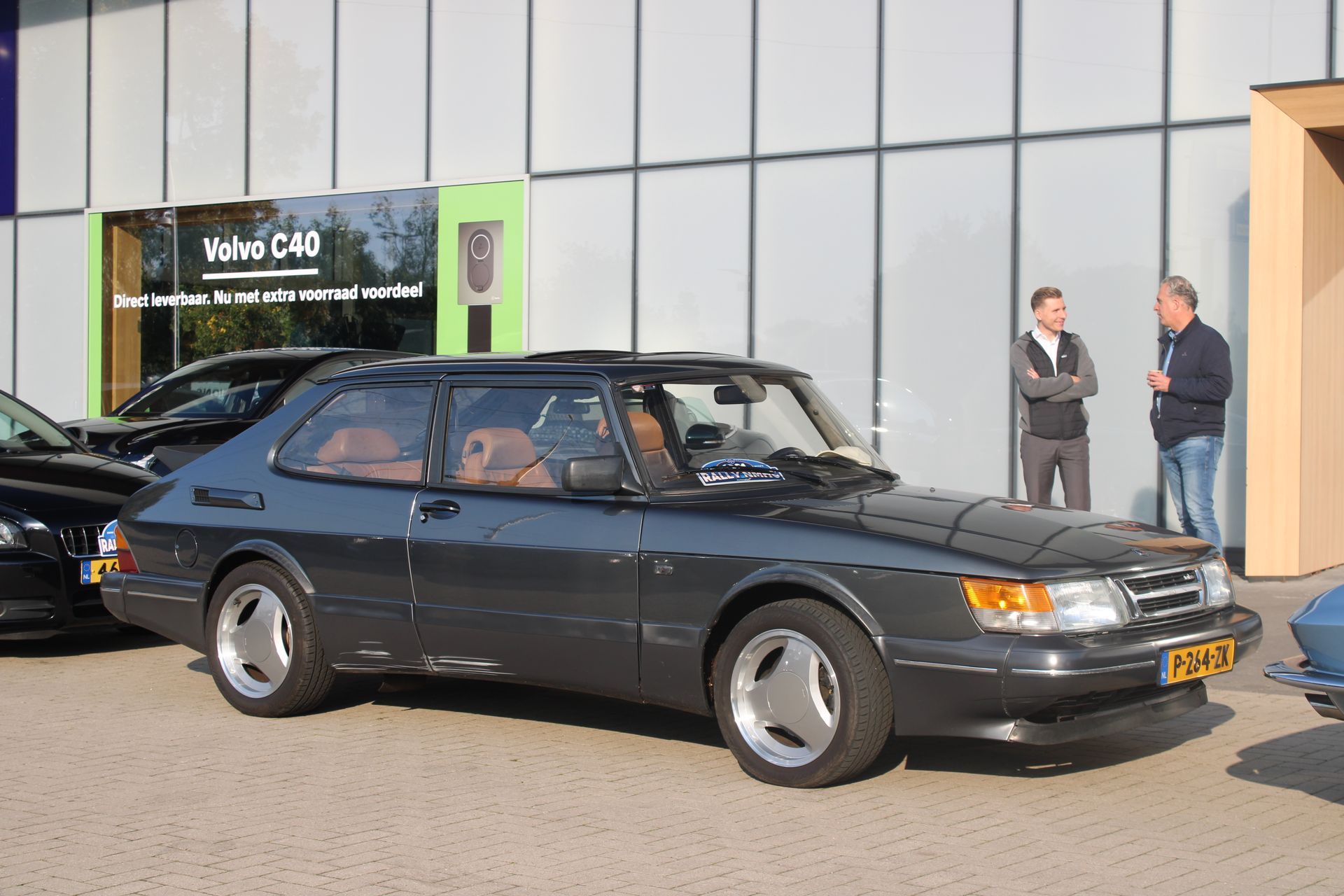 Een donkergrijze Saab 900 coupé staat geparkeerd voor een gebouw, met twee mannen ernaast.