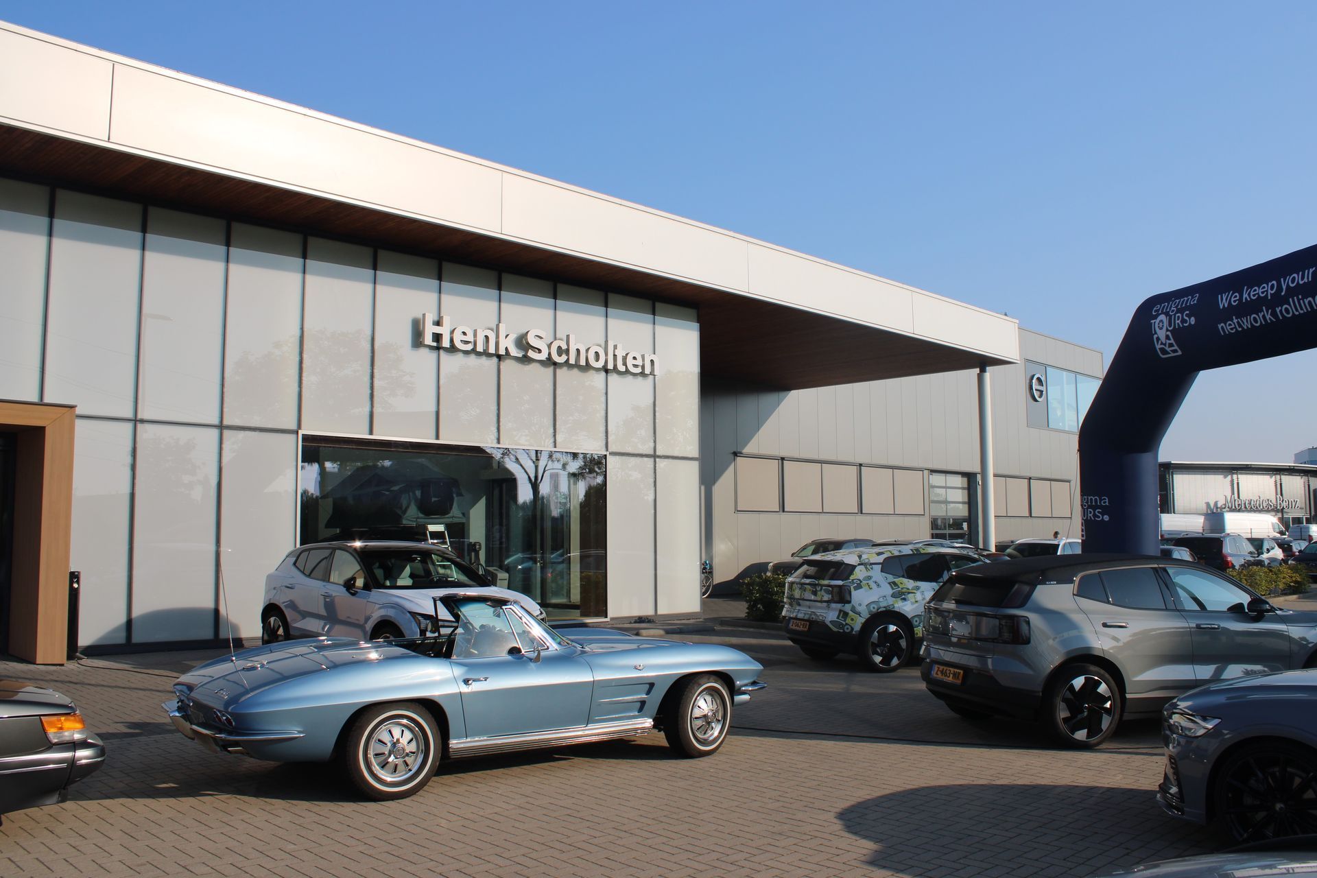 Een blauwe klassieke auto staat geparkeerd voor een autodealer, Henk Scholler. Andere auto's staan ​​op de parkeerplaats, de lucht is helder.