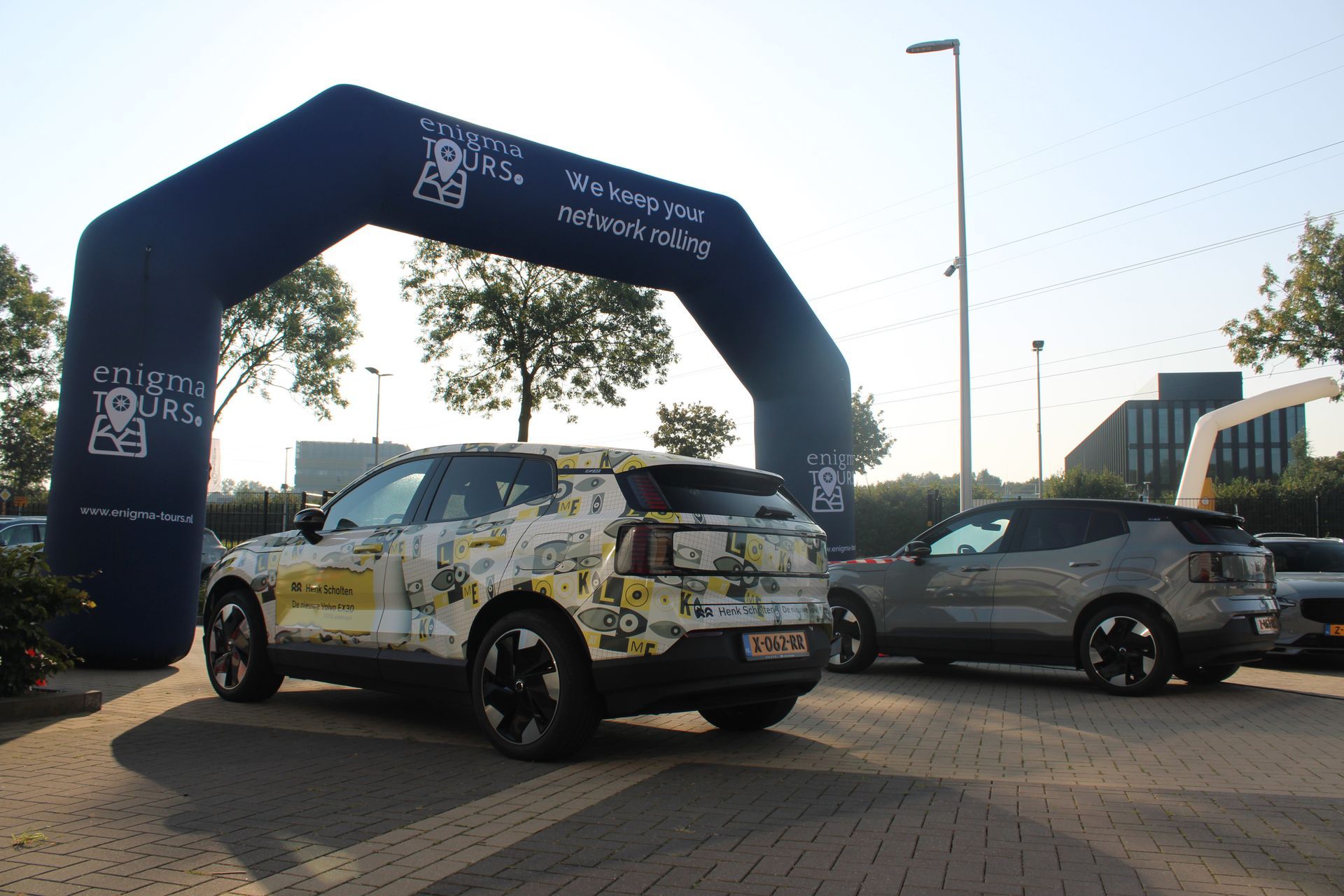Twee auto's onder een opblaasbare boog; de ene gecamoufleerd, de andere grijs. Buitenlocatie, logo zichtbaar.