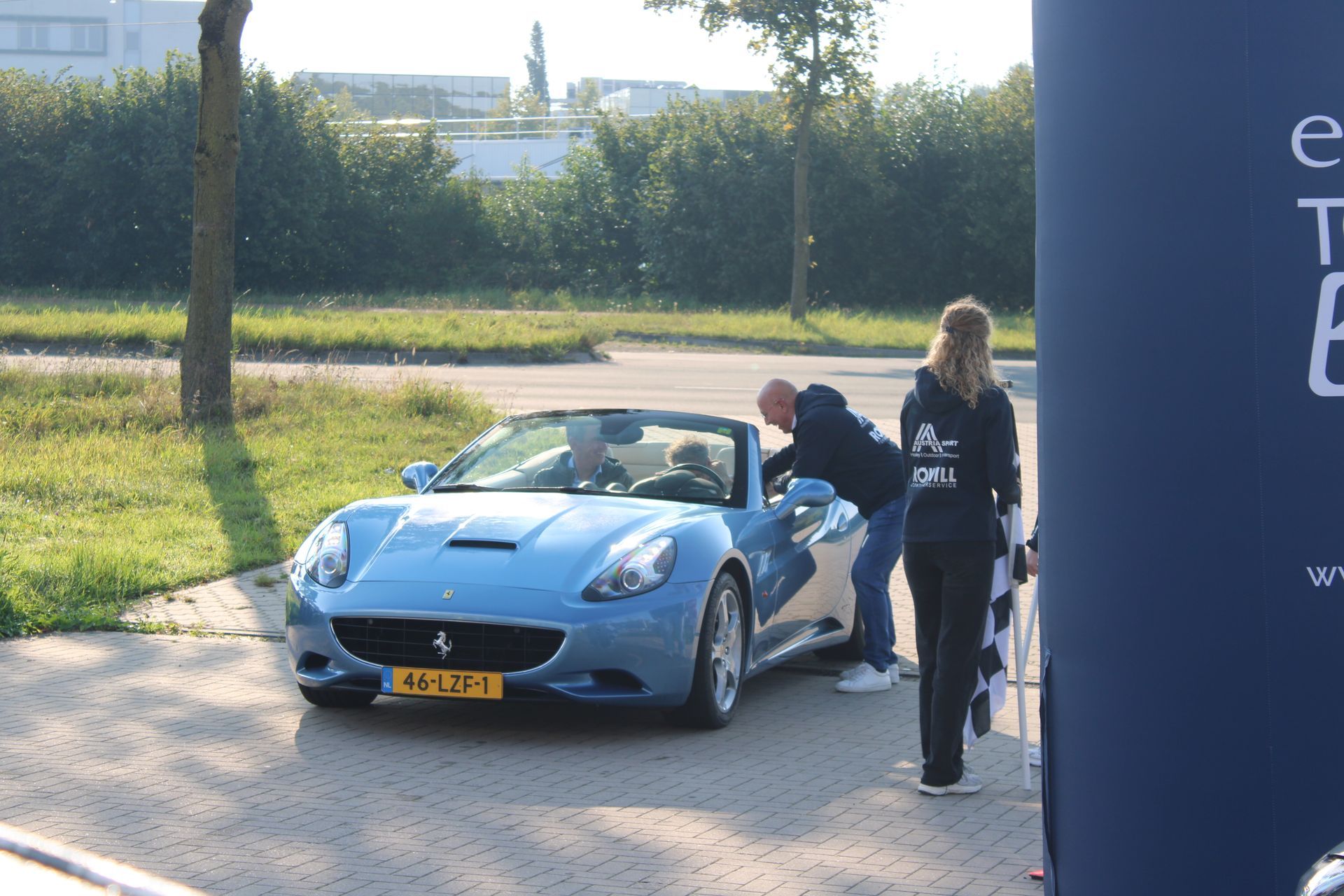 Lichtblauwe Ferrari cabriolet; twee mannen en een vrouw staan ​​erbij, mogelijk bij een evenement.