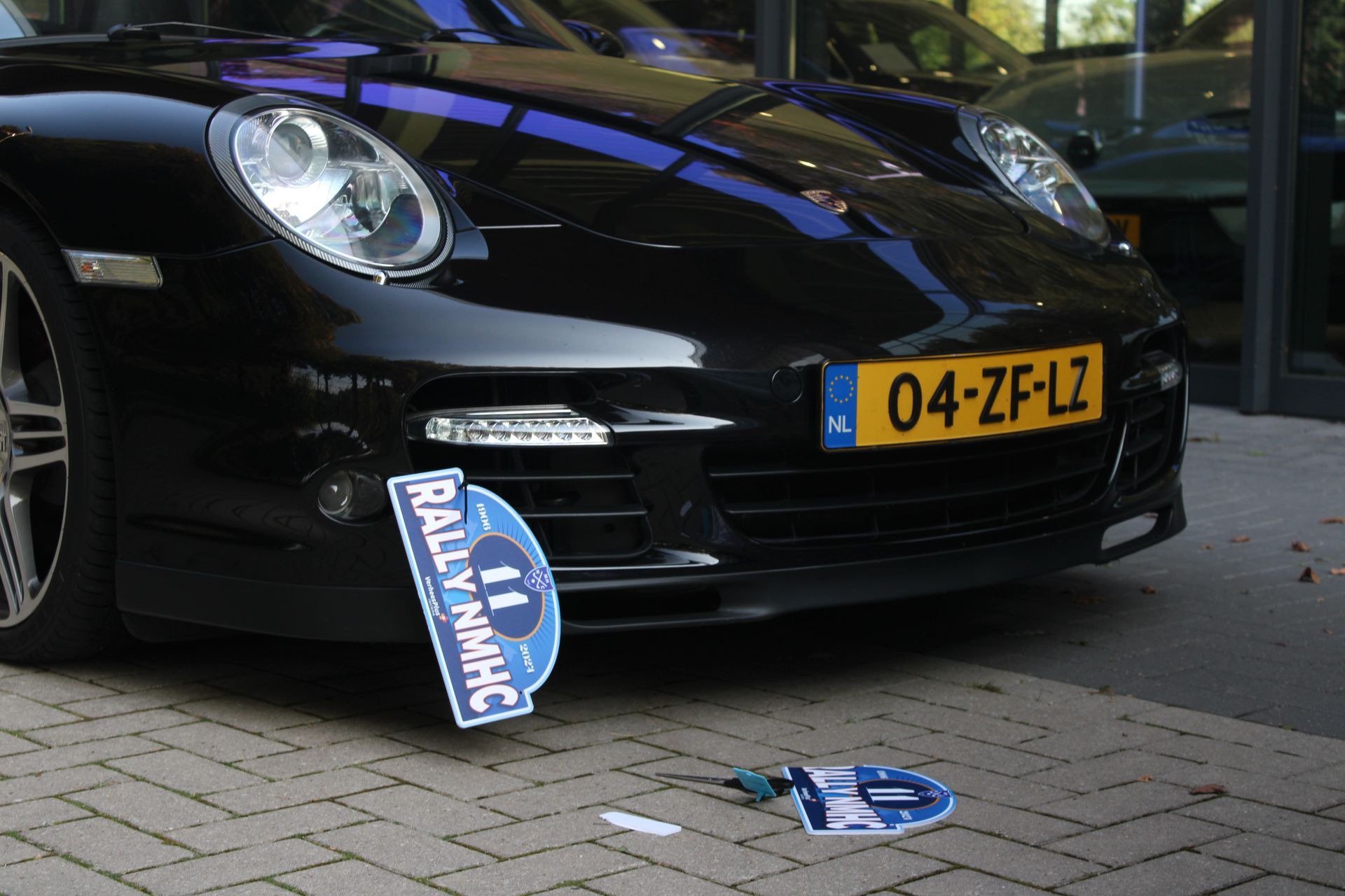 Zwarte Porsche met Nederlands kenteken en losgeraakte rallyplaat op bakstenen stoep.