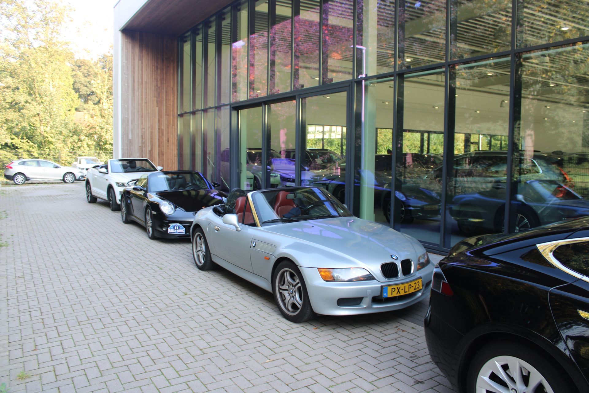 Een rij auto's staat geparkeerd voor een gebouw met glazen wanden; een zilverkleurige BMW cabriolet staat ervoor.