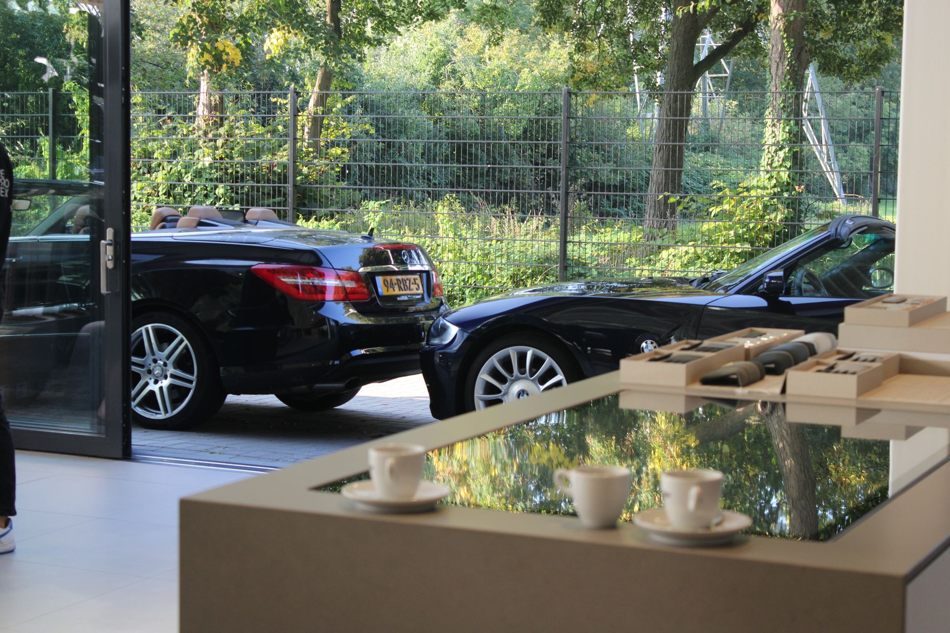 Twee zwarte cabriolets geparkeerd bij een open deuropening; interieur met een spiegelende tafel gedekt met kopjes.