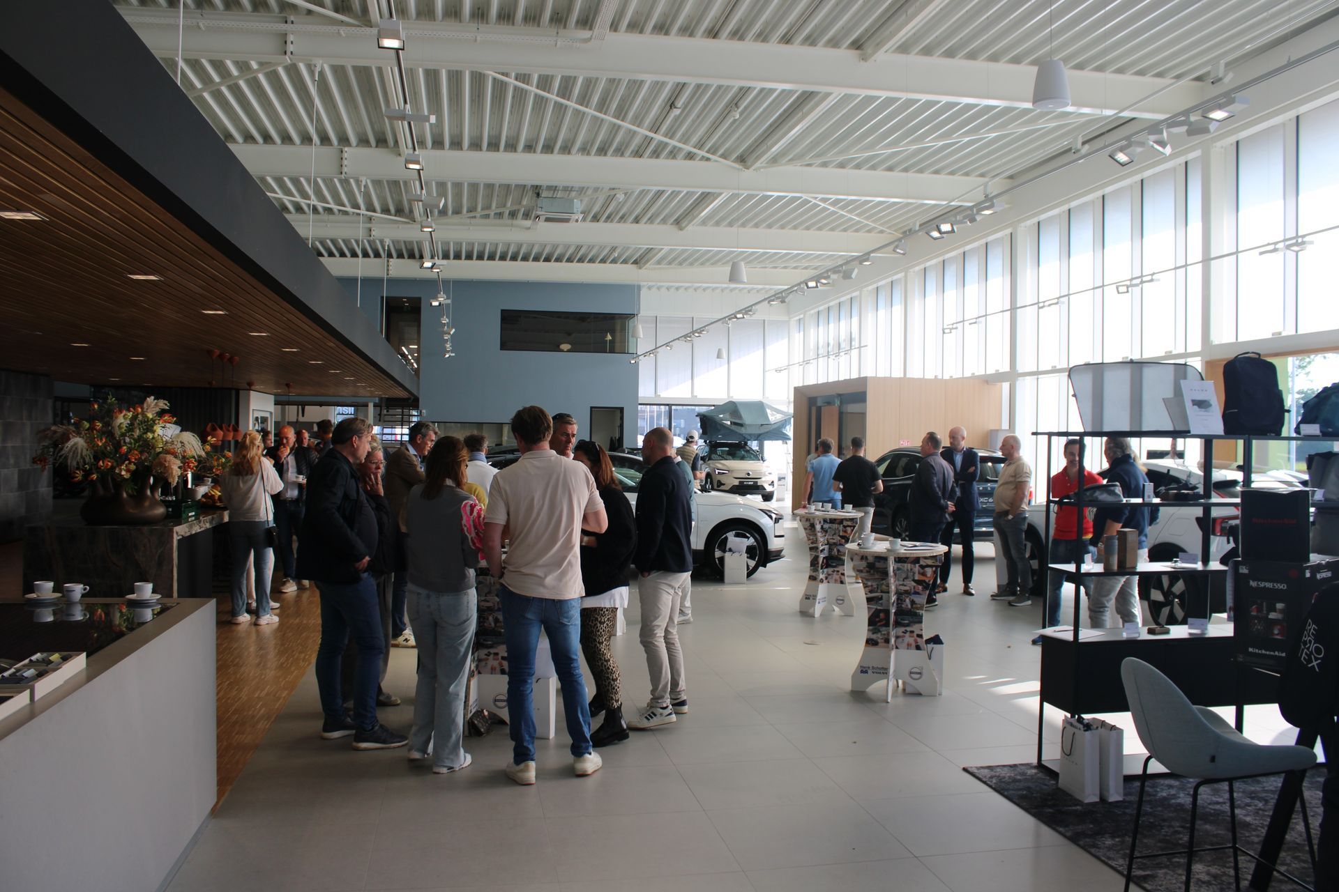 Mensen in een autoshowroom. Een witte auto is zichtbaar, die naast een tafel met andere artikelen staat.