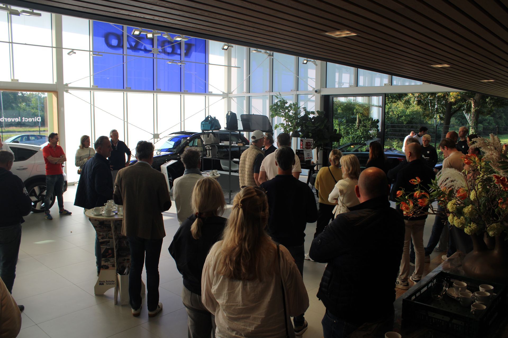 Een groep mensen in een lichte ruimte, waarschijnlijk een showroom. Auto's zijn zichtbaar, een blauwe muur met een logo op de achtergrond.