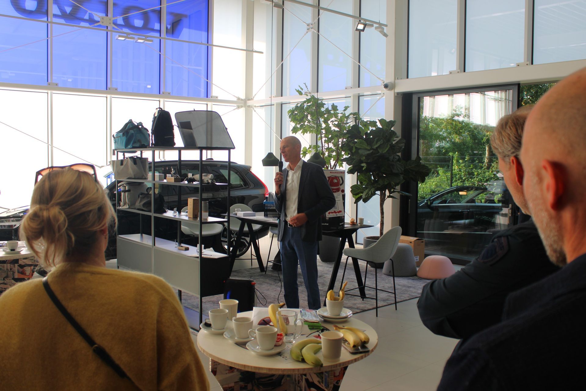 Een man spreekt op een Volvo-evenement, staand bij een tafel met hapjes en drankjes. Mensen luisteren, auto's rijden op de achtergrond.
