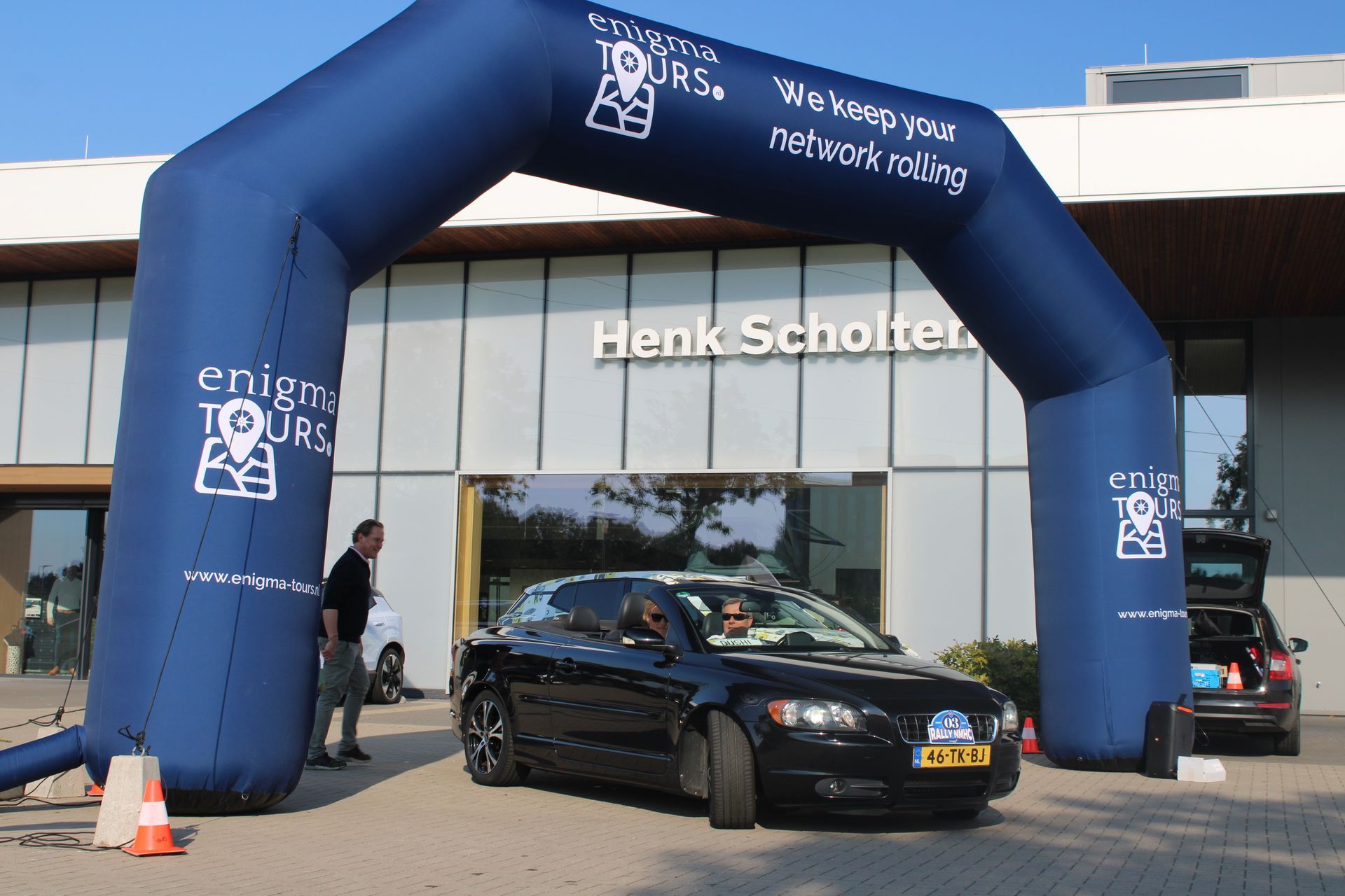 Een zwarte auto rijdt onder een opblaasbare boog door tijdens een evenement, met een gebouw op de achtergrond.