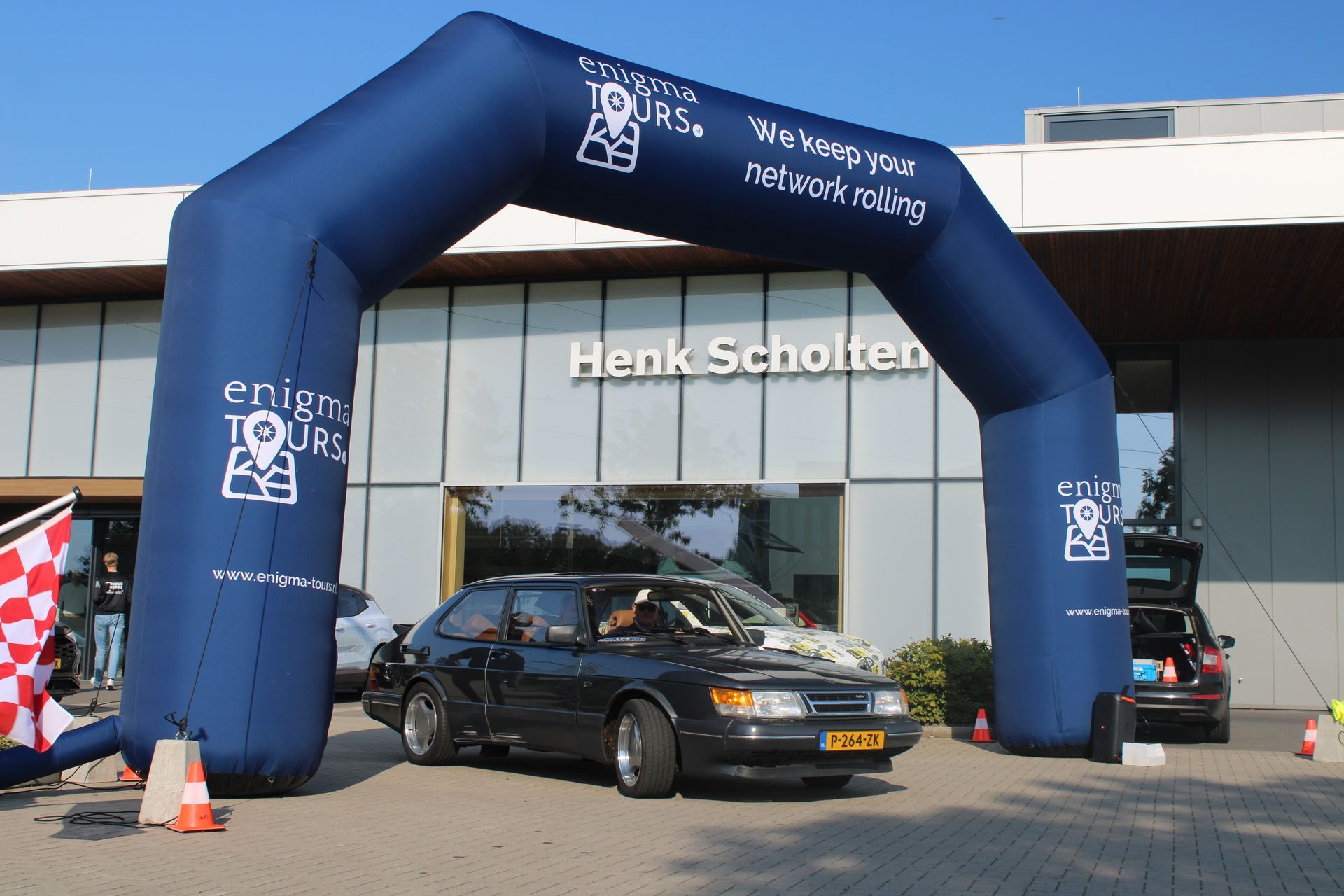 Een donkere auto rijdt onder een blauwe opblaasbare boog door, met op de achtergrond het Henk Scholten-gebouw.