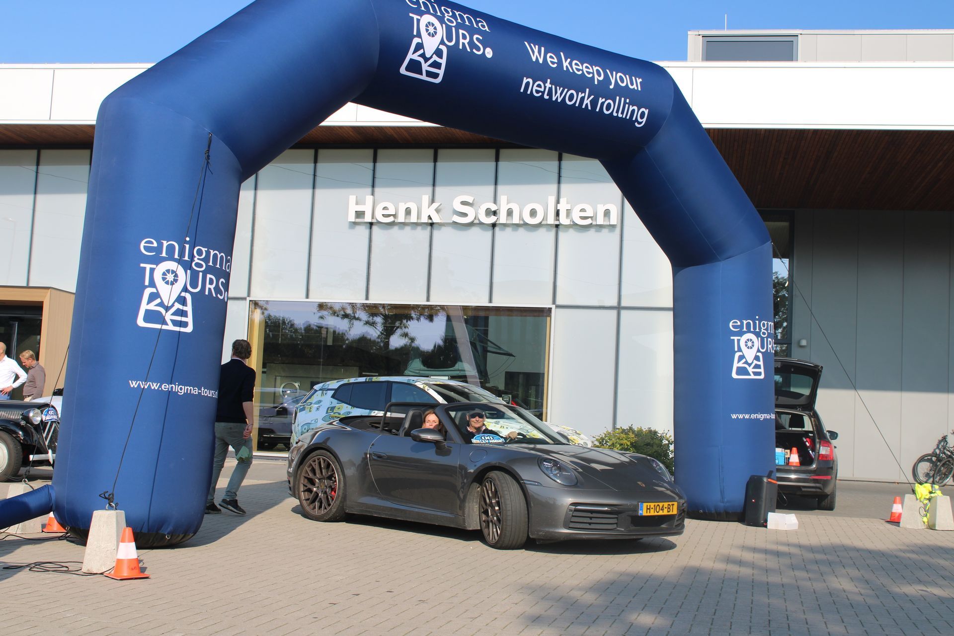 Een grijze sportwagen rijdt onder een blauwe opblaasbare boog door, voor het gebouw van Henk Scholten.