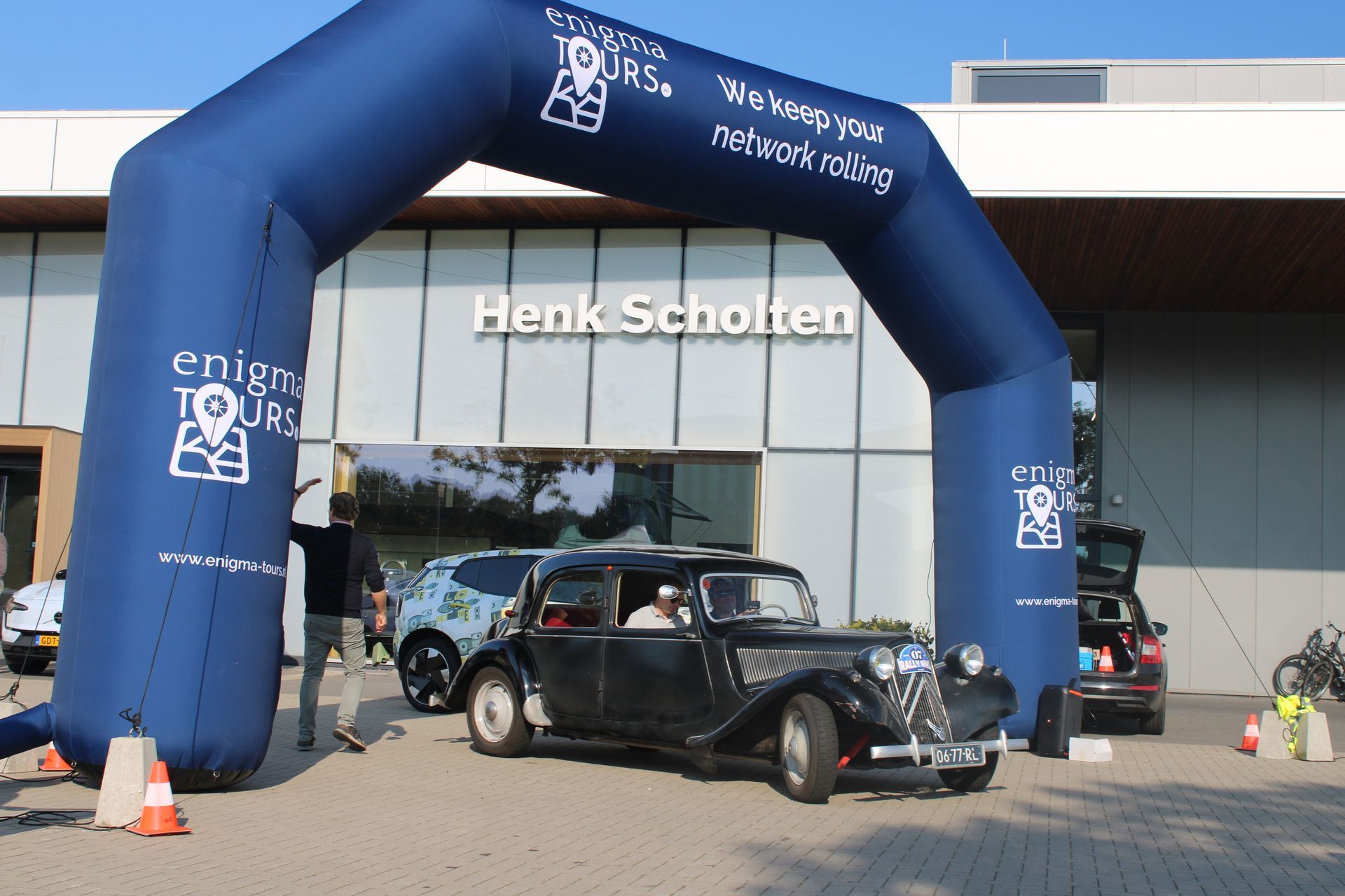 Een zwarte oldtimer rijdt onder een blauwe boog door, met de opschriften 