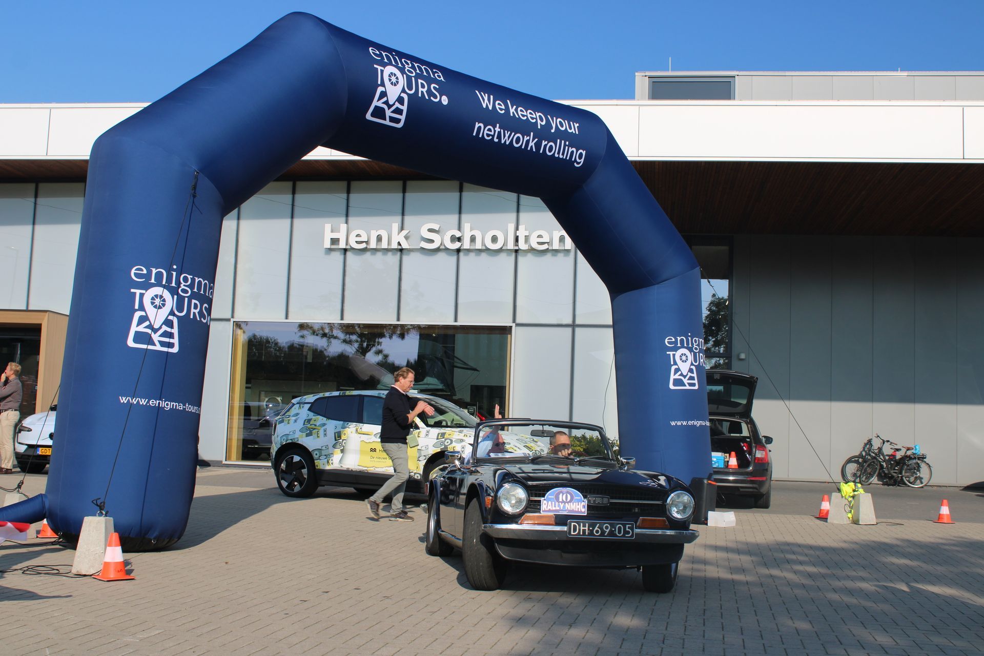 Blauwe boog boven auto's; man loopt langs een zwarte cabriolet. 
