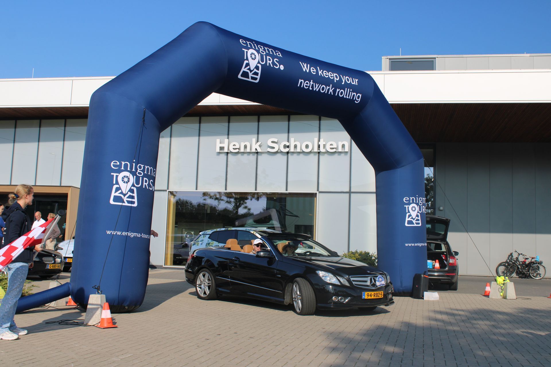 Een zwarte auto rijdt onder een blauwe opblaasbare boog door, met het Henk Scholten-gebouw op de achtergrond.