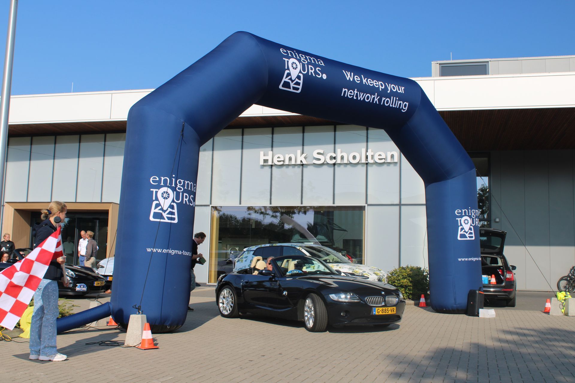 Een zwarte auto rijdt onder een blauwe opblaasbare boog door, met het Henk Schotten-gebouw op de achtergrond. Iemand zwaait met een geblokte vlag.