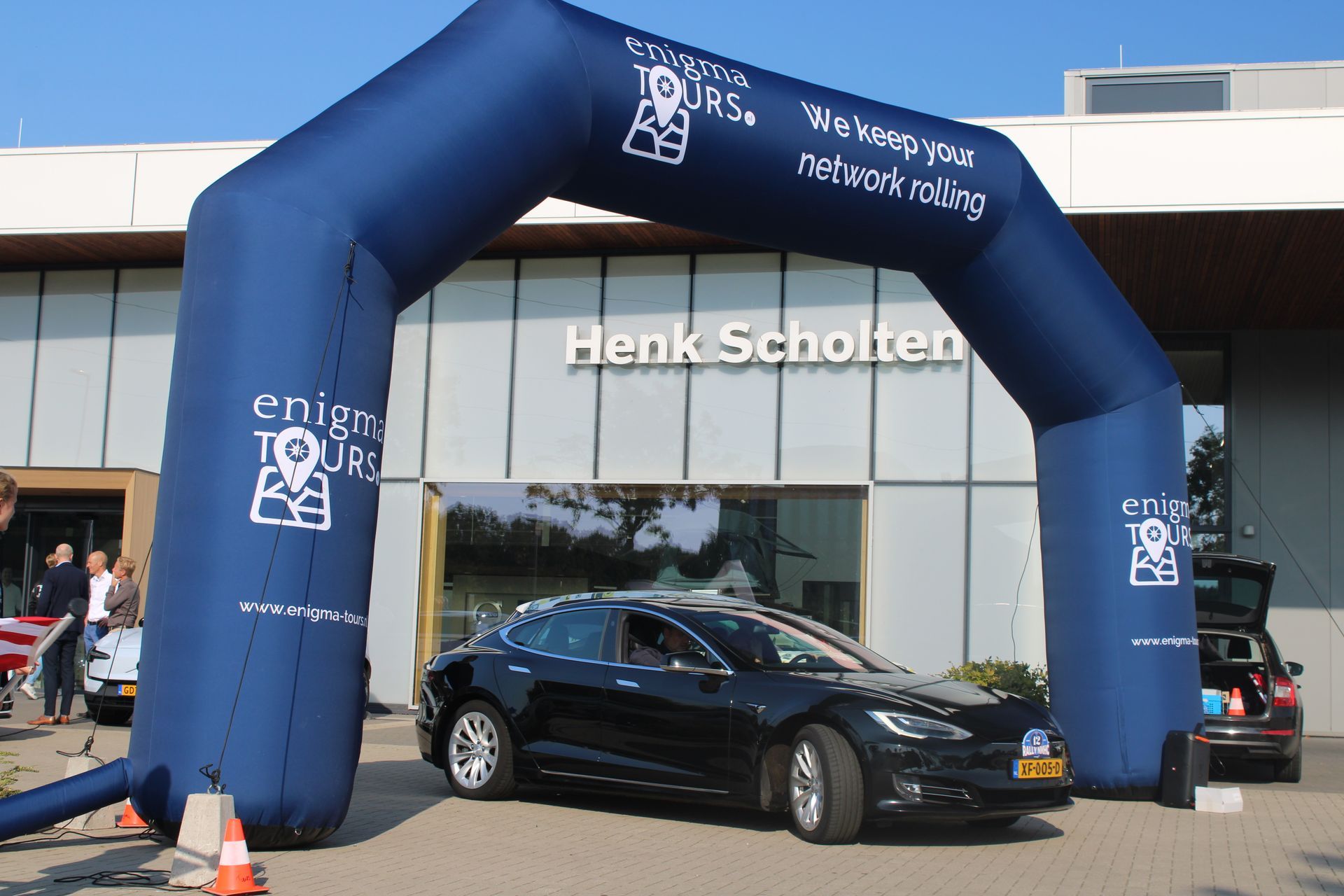 Een zwarte auto rijdt onder een blauwe opblaasbare boog door, met het Henk Scholten-gebouw op de achtergrond.