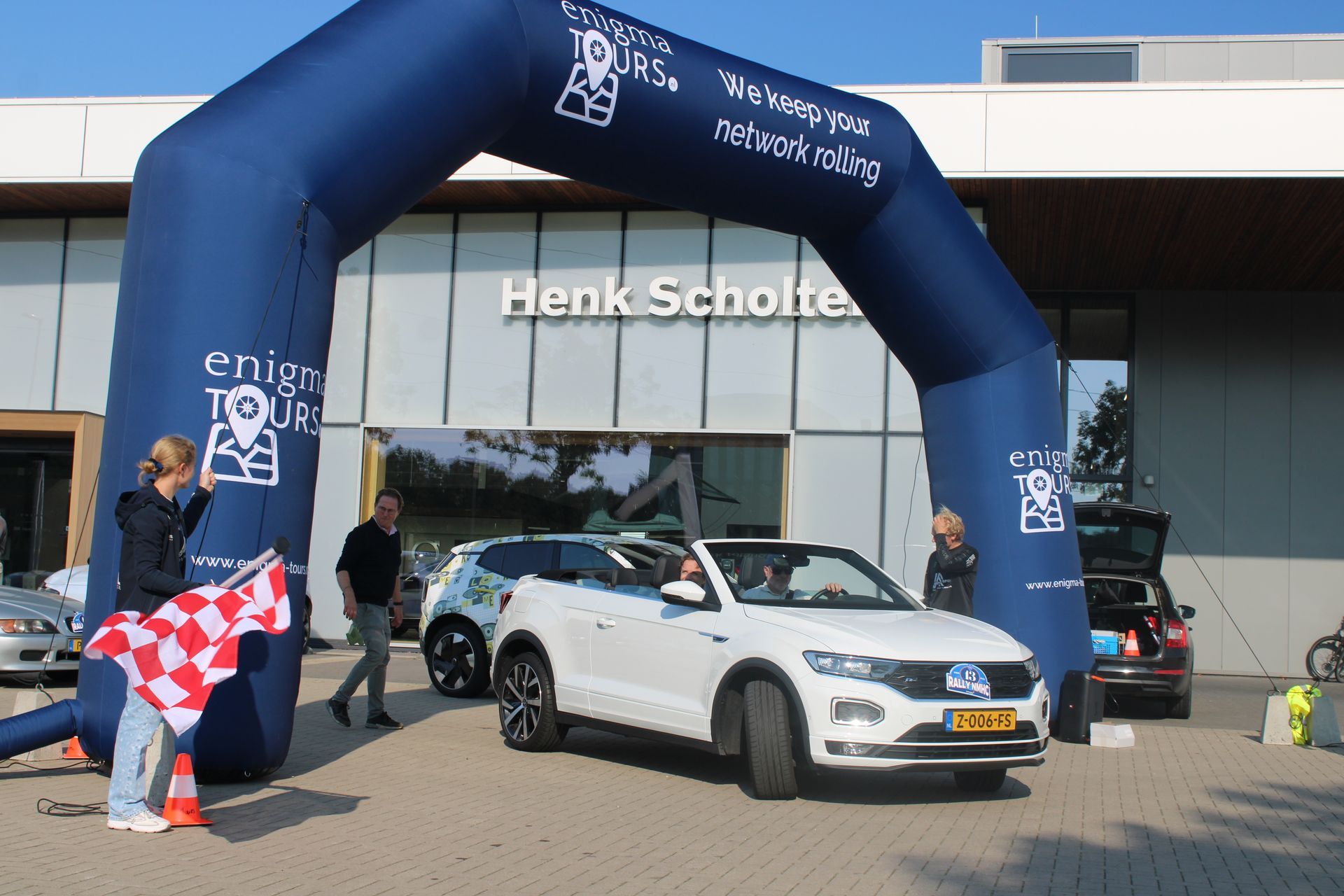 Een witte auto rijdt onder een blauwe opblaasbare boog door. Mensen kijken toe en houden een geblokte vlag vast. Op de achtergrond is een gebouw te zien.