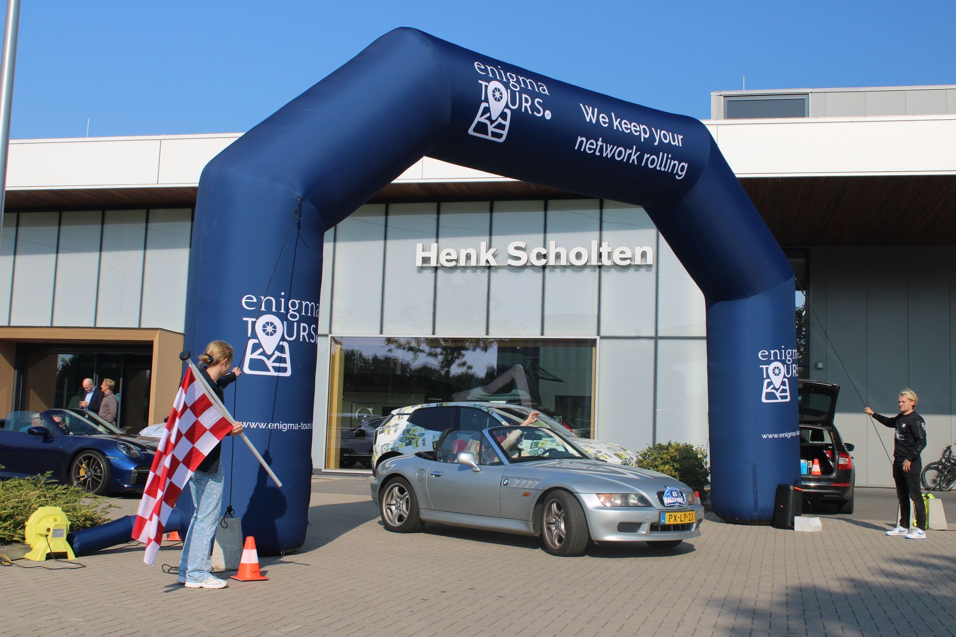 Een zilverkleurige auto passeert de finishboog bij de Henk Scholten-dealer. Iemand zwaait met de geblokte vlag.