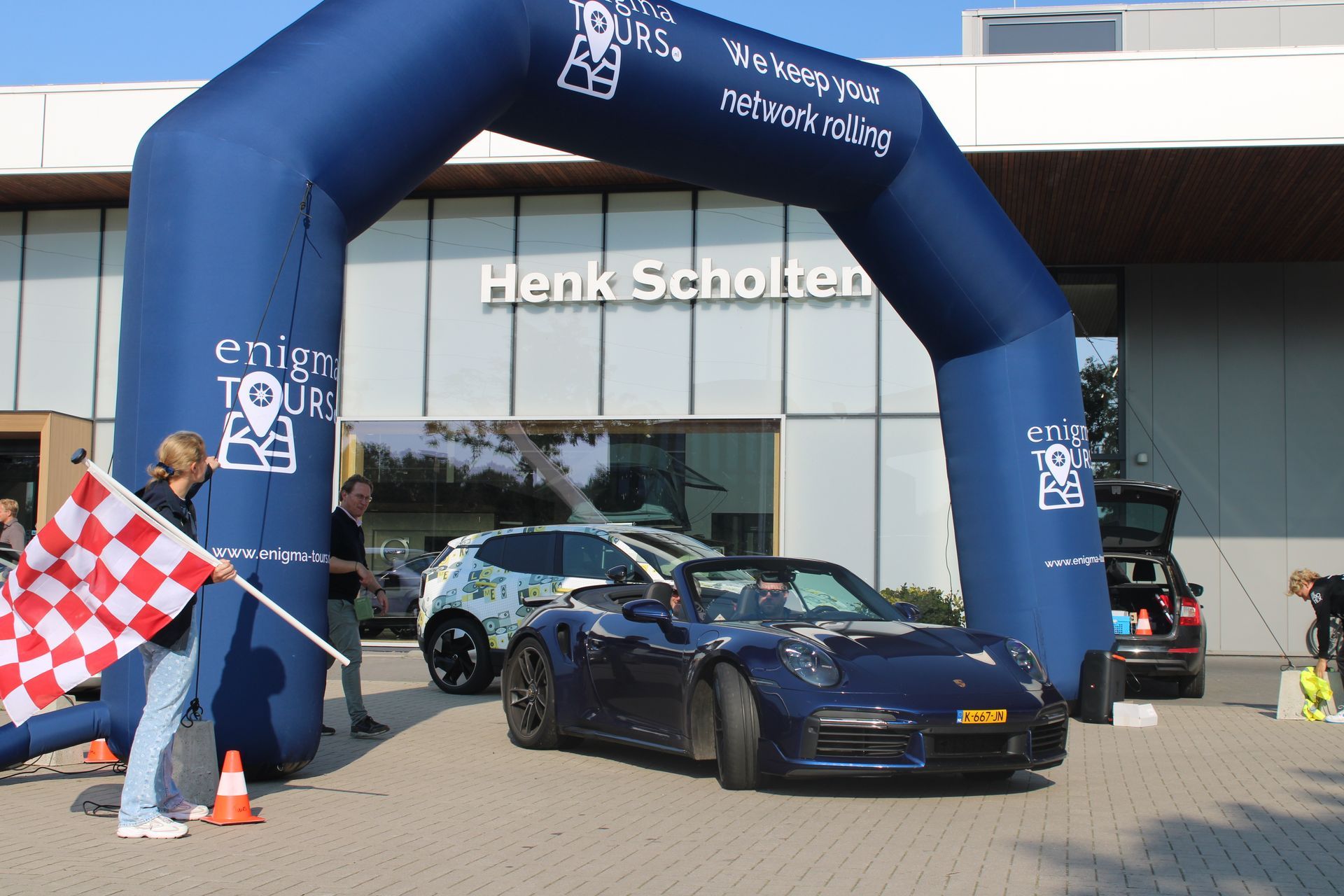 Een blauwe Porsche cabriolet passeert de finishlijn bij een evenement, met het Henk Scholten-gebouw op de achtergrond.