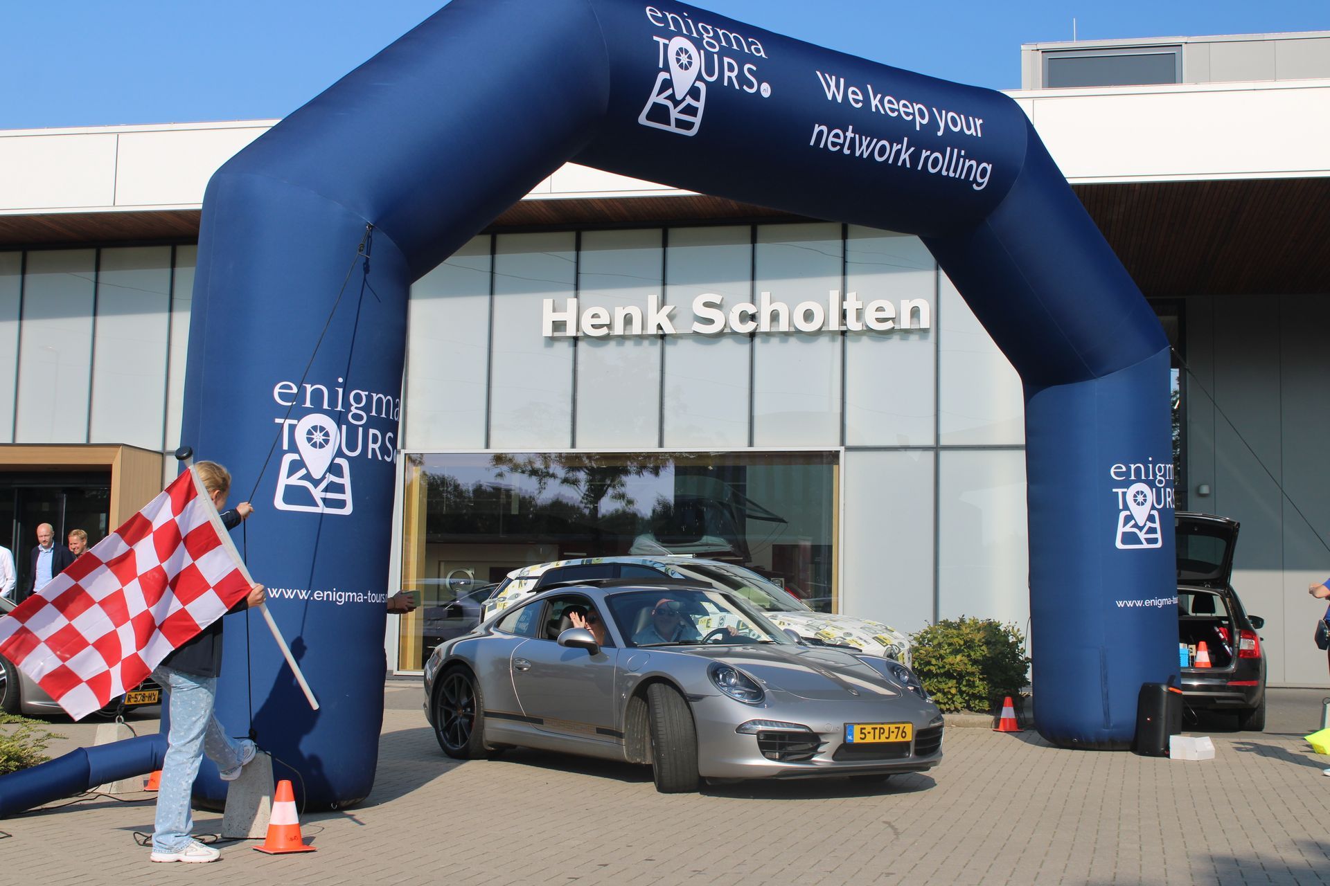 Een zilverkleurige auto rijdt door de finishboog van Enigma Tours; iemand zwaait met een geblokte vlag.