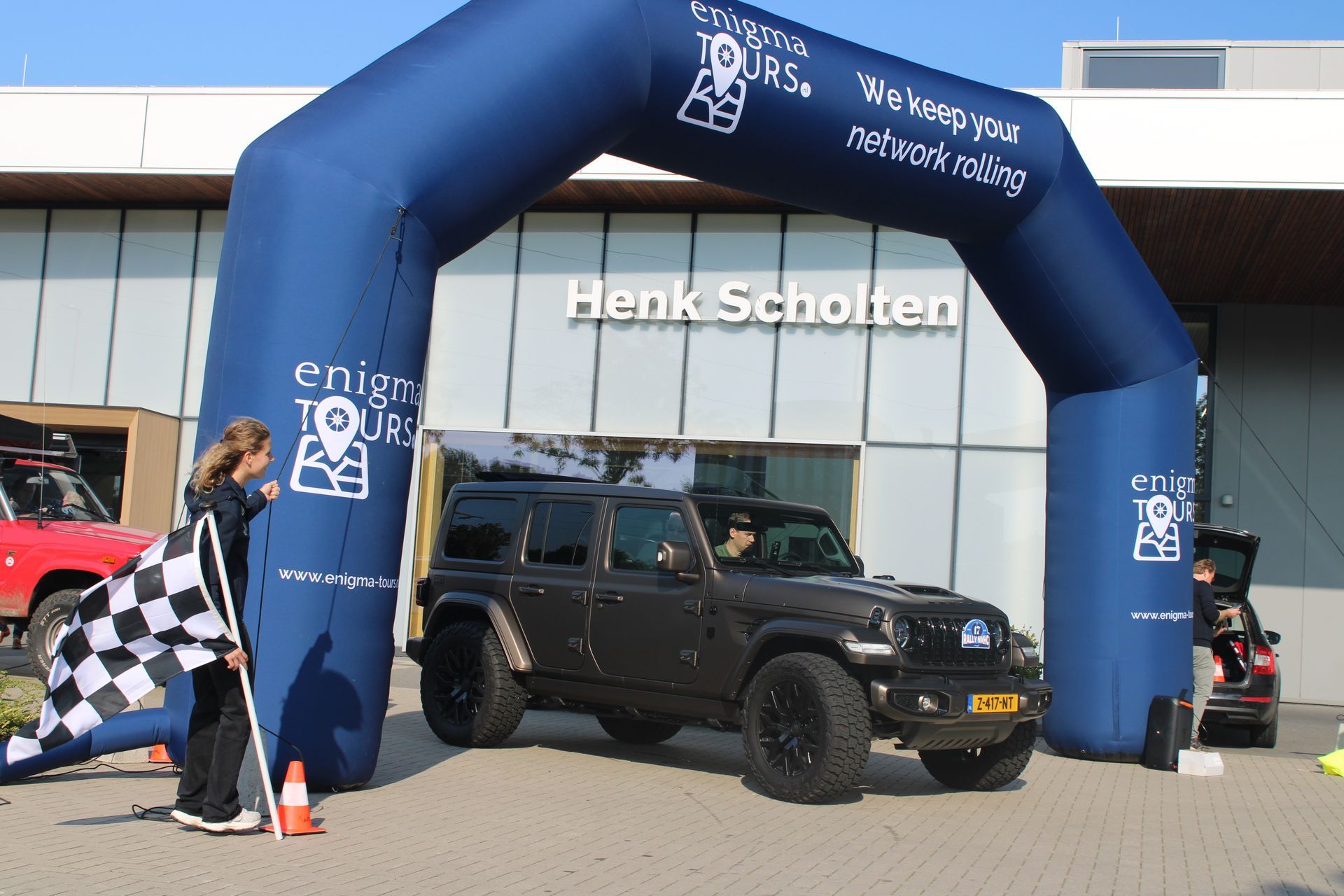 Een donkere SUV rijdt onder een blauwe opblaasbare boog door, het Henk Scholten-gebouw, met een wapperende geblokte vlag.