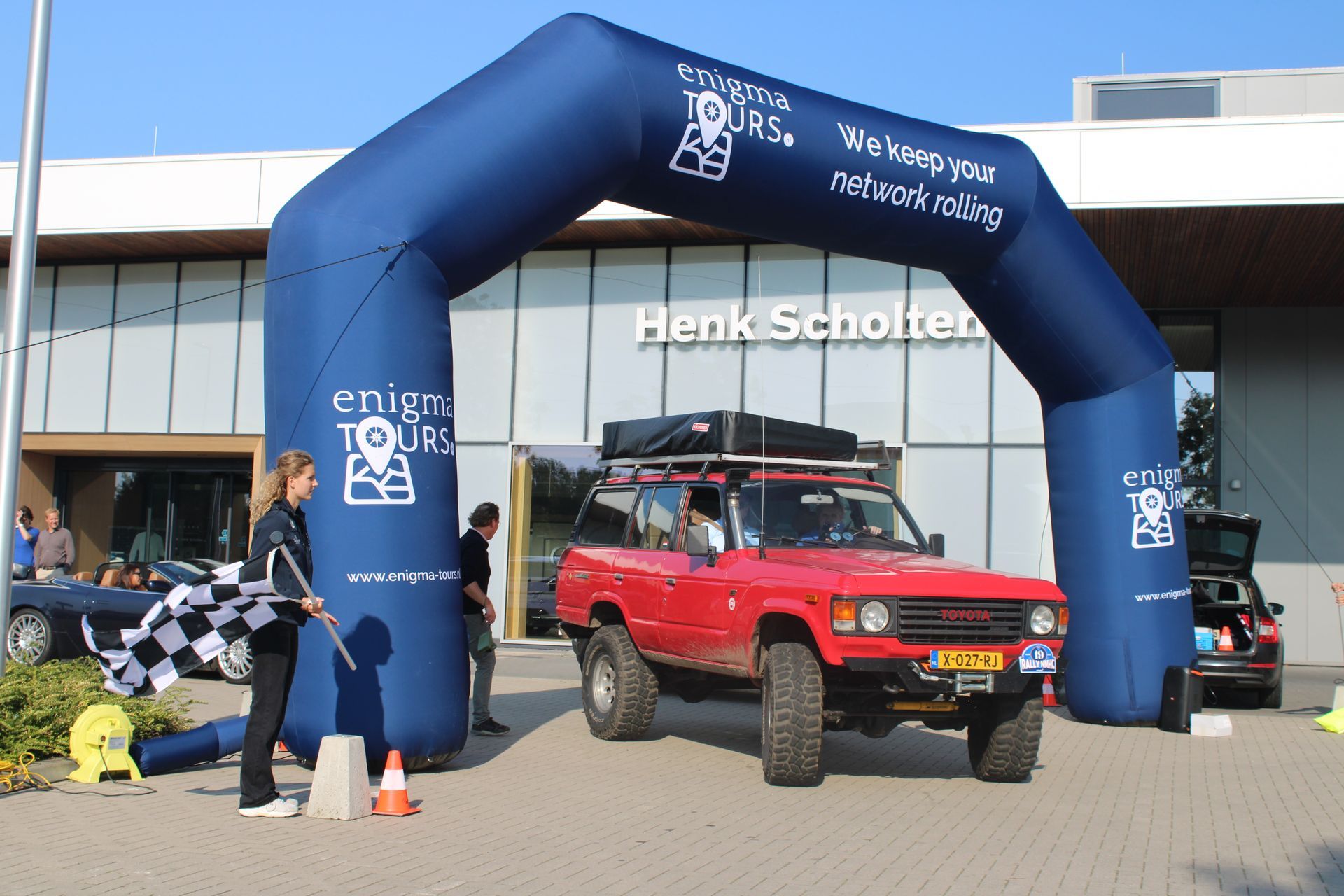 Een rode SUV rijdt onder een blauwe opblaasbare boog door, terwijl een vrouw met een geblokte vlag zwaait.