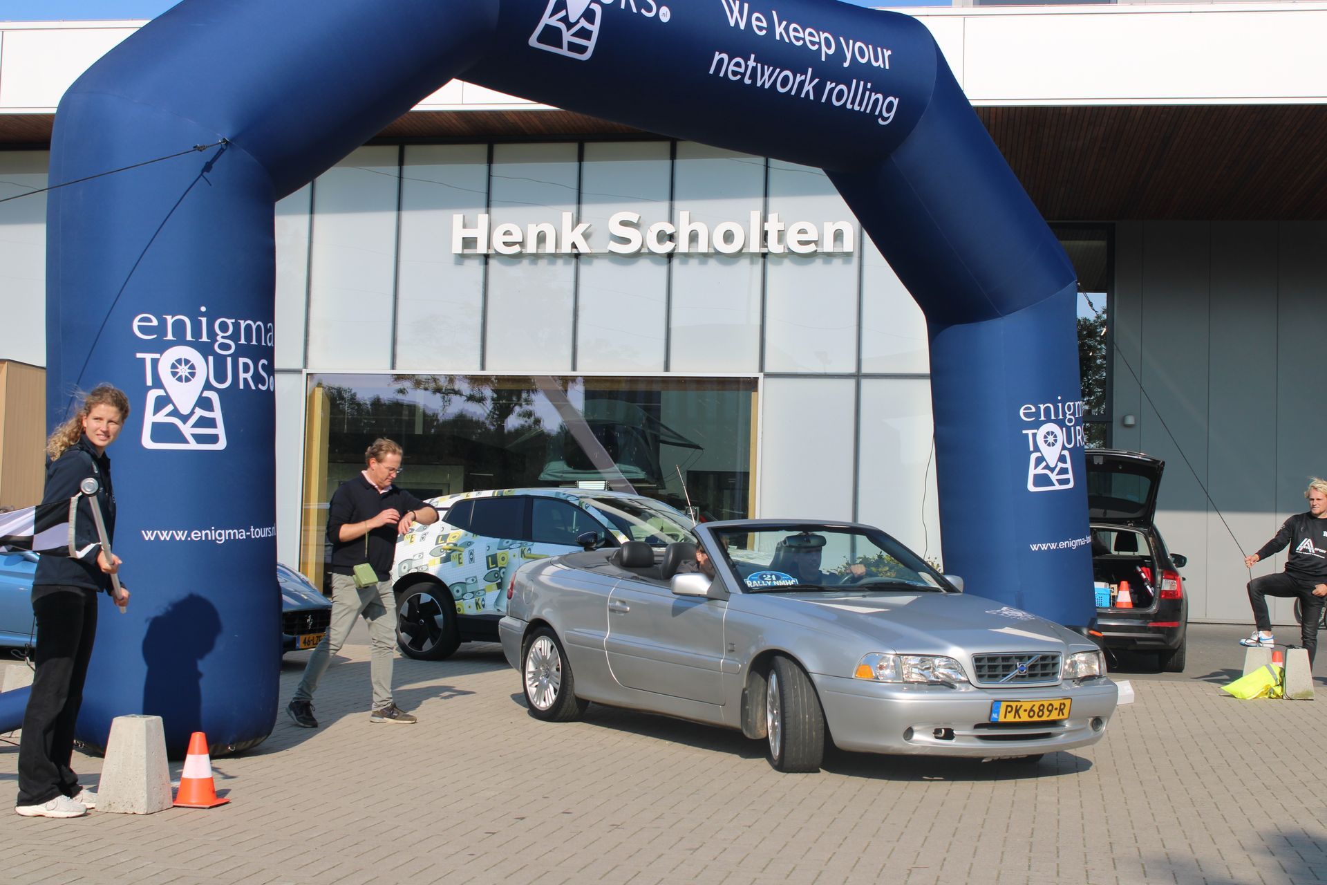 Een zilverkleurige cabriolet staat geparkeerd onder een blauwe opblaasbare boog met het opschrift 