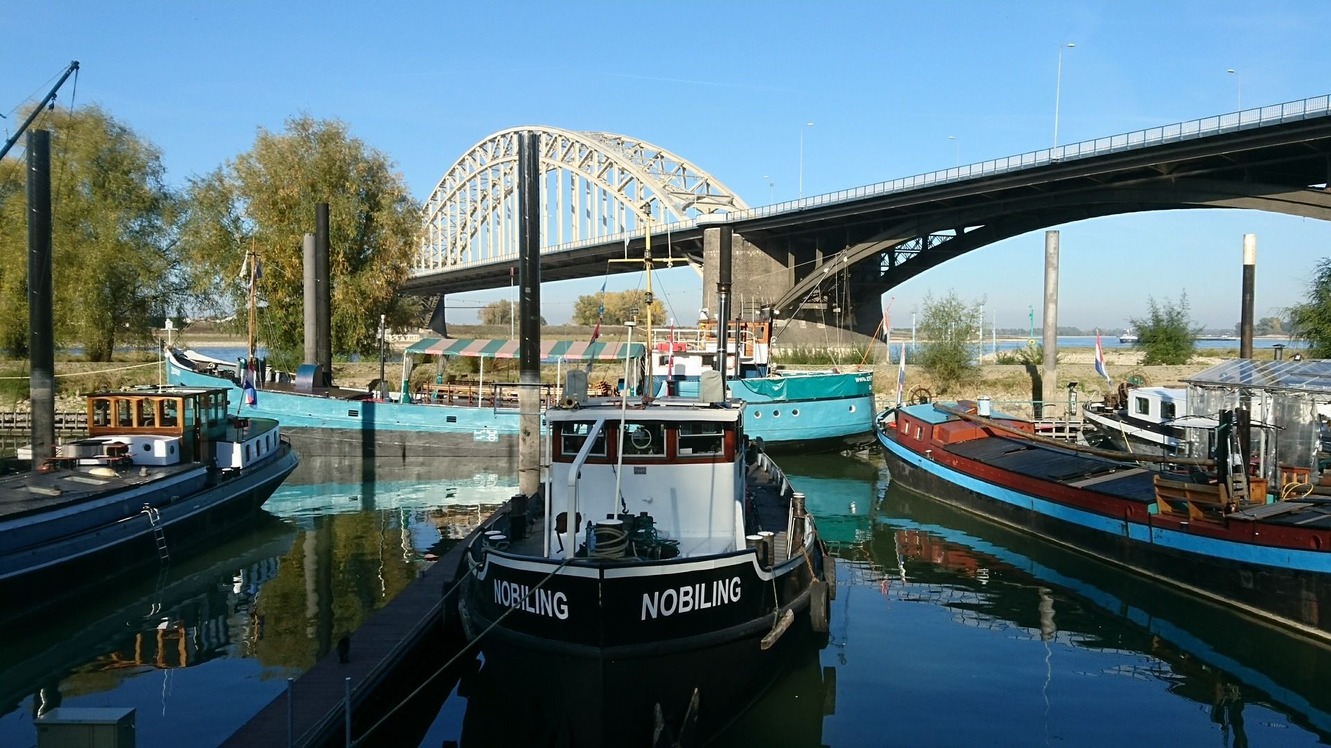 Boten liggen aangemeerd in een haven onder een grote brug. Het water weerspiegelt de schepen en de lucht.