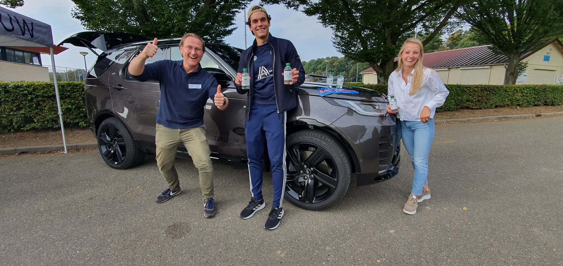 Drie mensen poseren bij een grijze SUV. Twee mannen maken handgebaren, een vrouw glimlacht. Buiten op het asfalt.