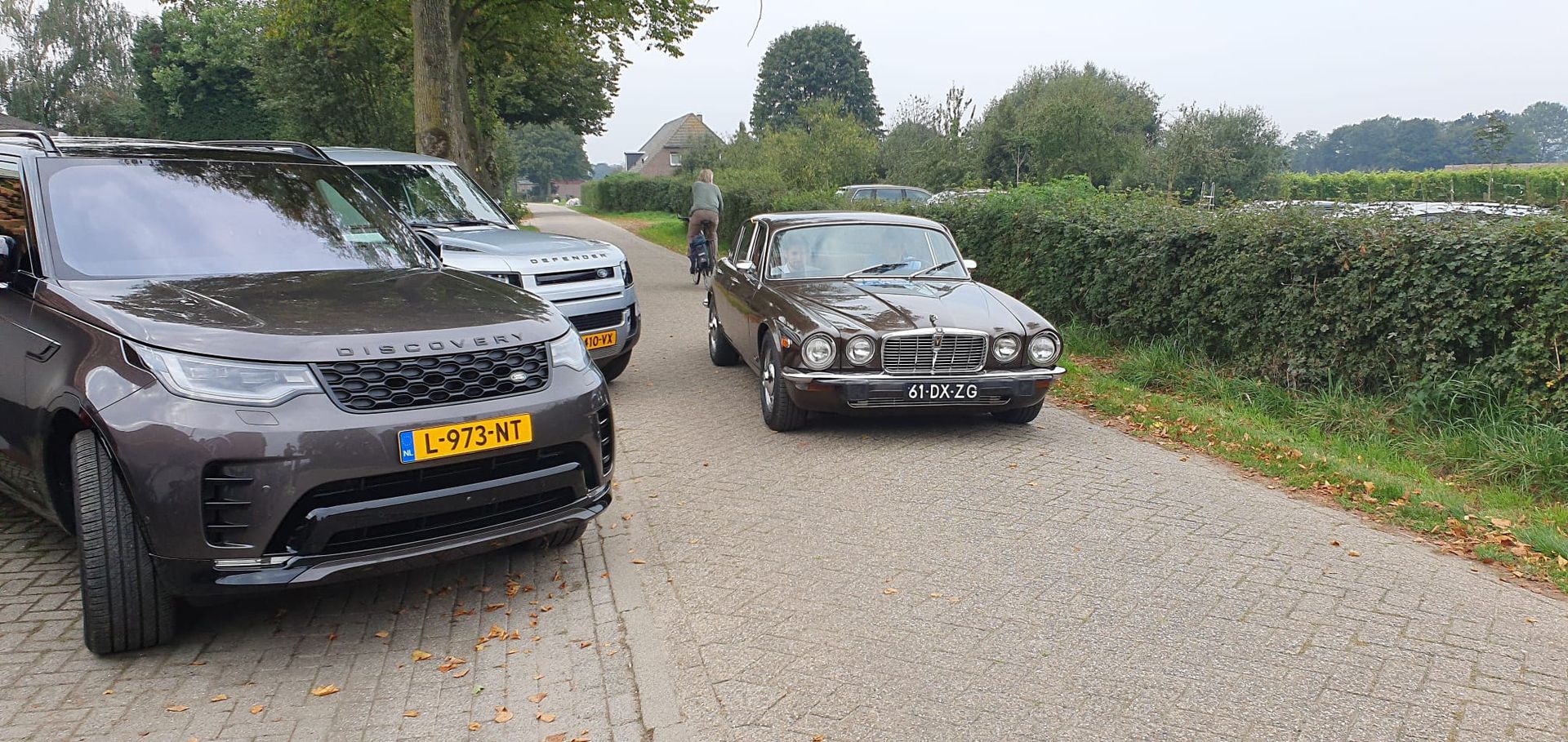 Drie auto's staan ​​geparkeerd op een grindweg, één is een bruine klassieke auto, twee zijn moderne SUV's.