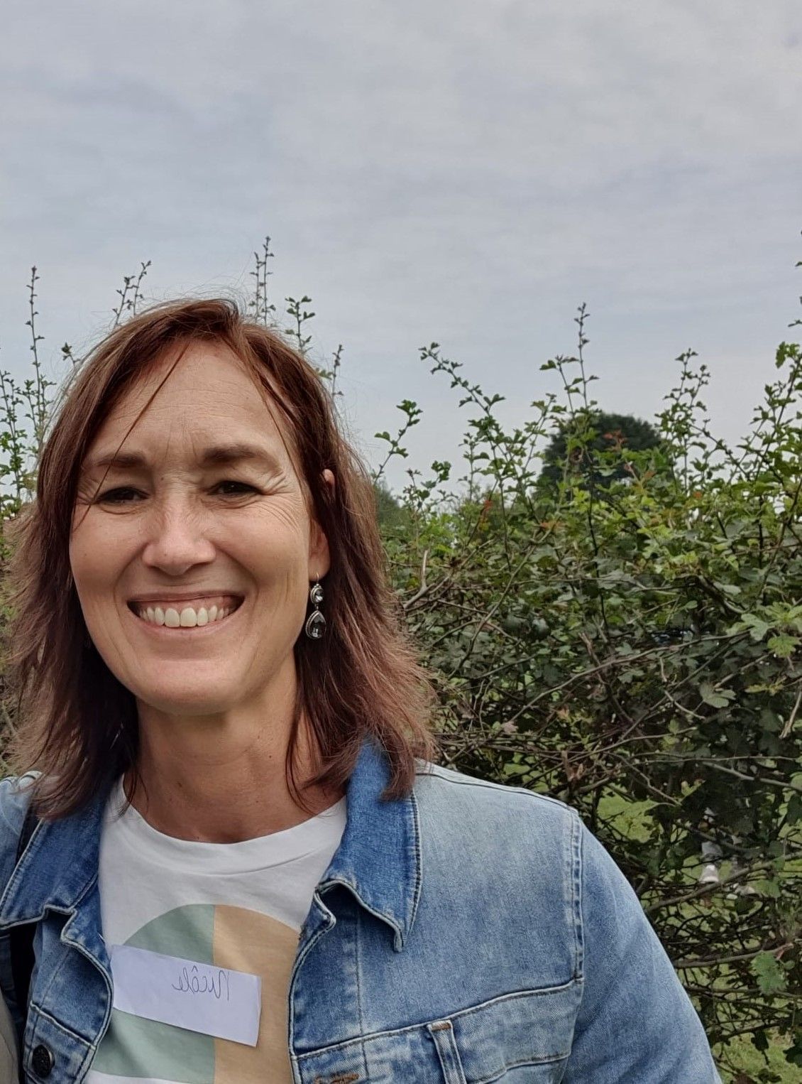 Een lachende vrouw, gekleed in een spijkerjasje, staat buiten bij groene struiken en bomen.