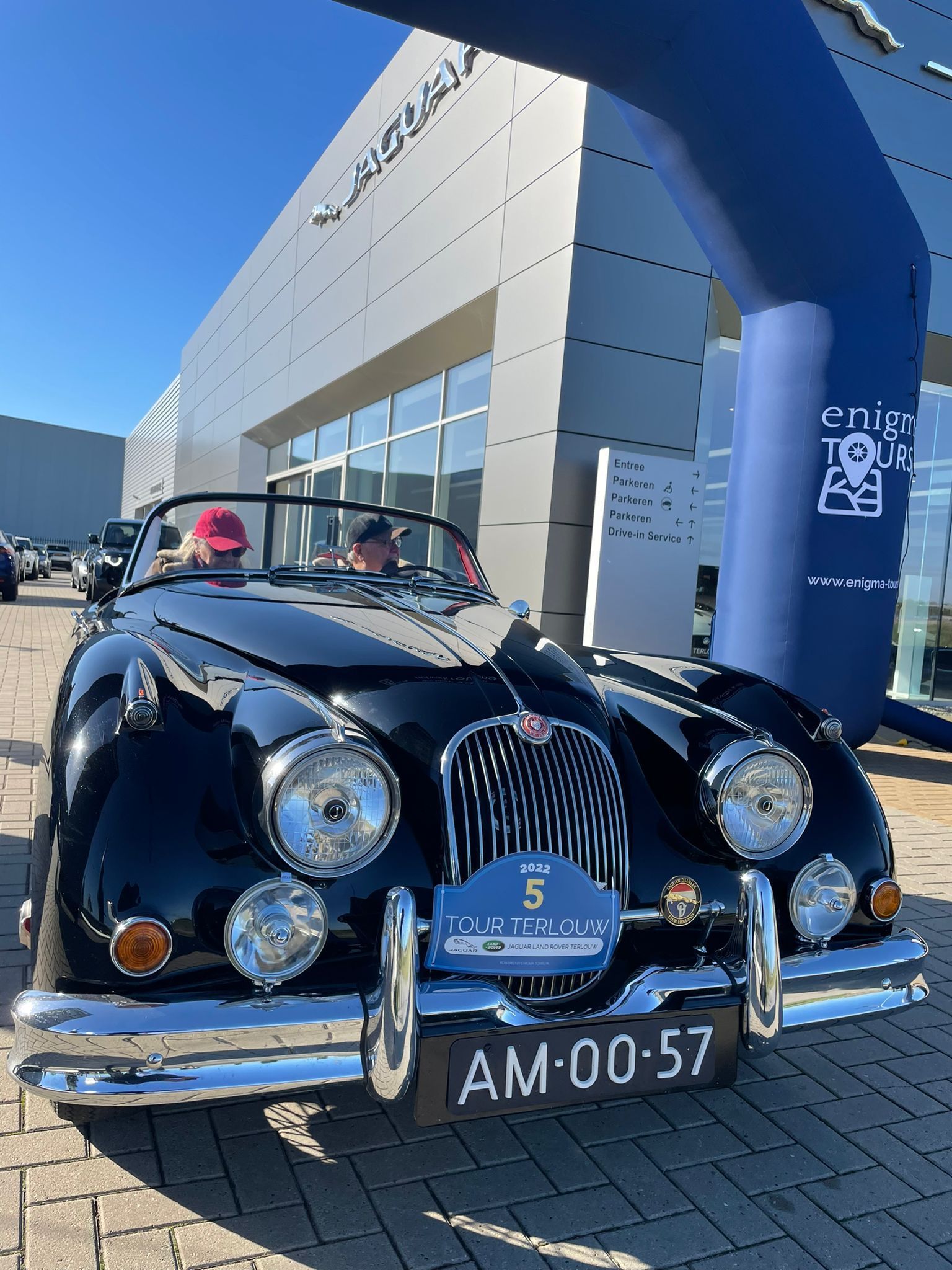 Een zwarte vintage Jaguar cabriolet staat voor een Jaguar-dealer; een blauwe boog is zichtbaar.