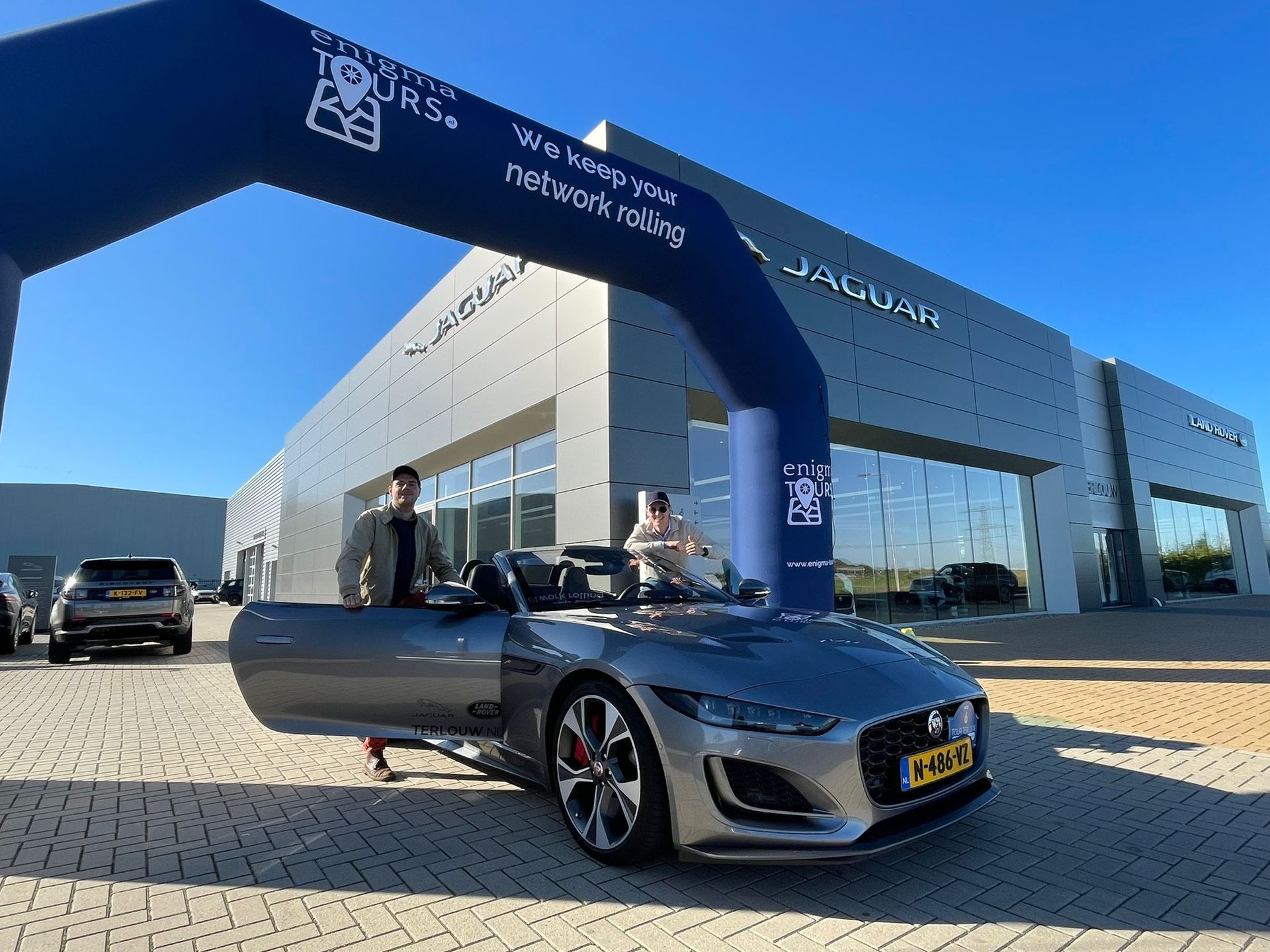 Twee mensen in een grijze Jaguar cabriolet voor een autodealer, onder een blauwe boog.