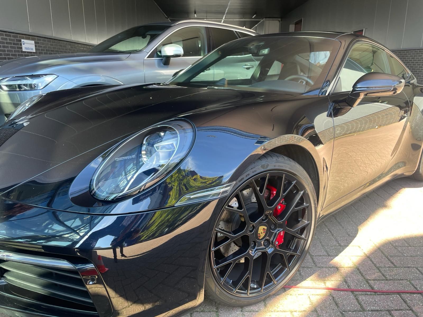 Een zwarte Porsche sportwagen met rode remklauwen staat geparkeerd onder een afdak; op de achtergrond is nog een auto te zien.
