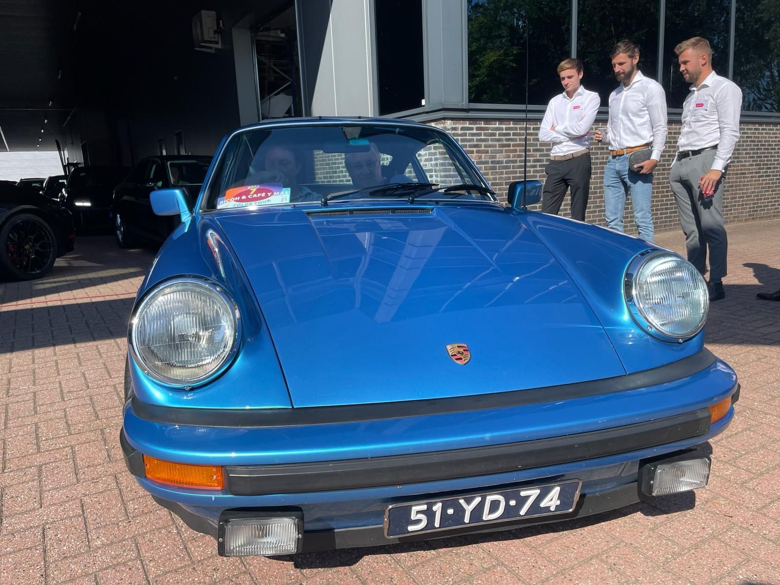 Een blauwe klassieke Porsche met drie mensen in witte overhemden en een bakstenen gebouw op de achtergrond.