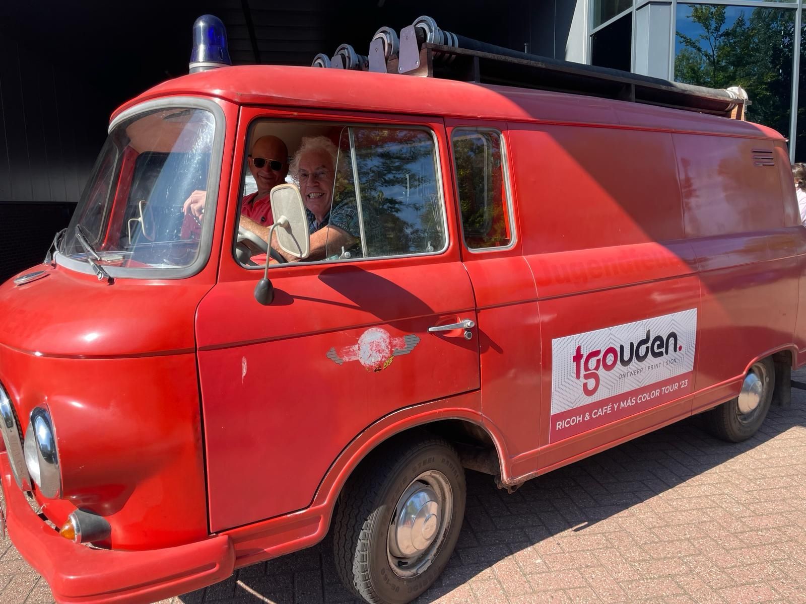 Rode oldtimerbus met twee inzittenden, geparkeerd. Op de zijkant van de bus staat het 