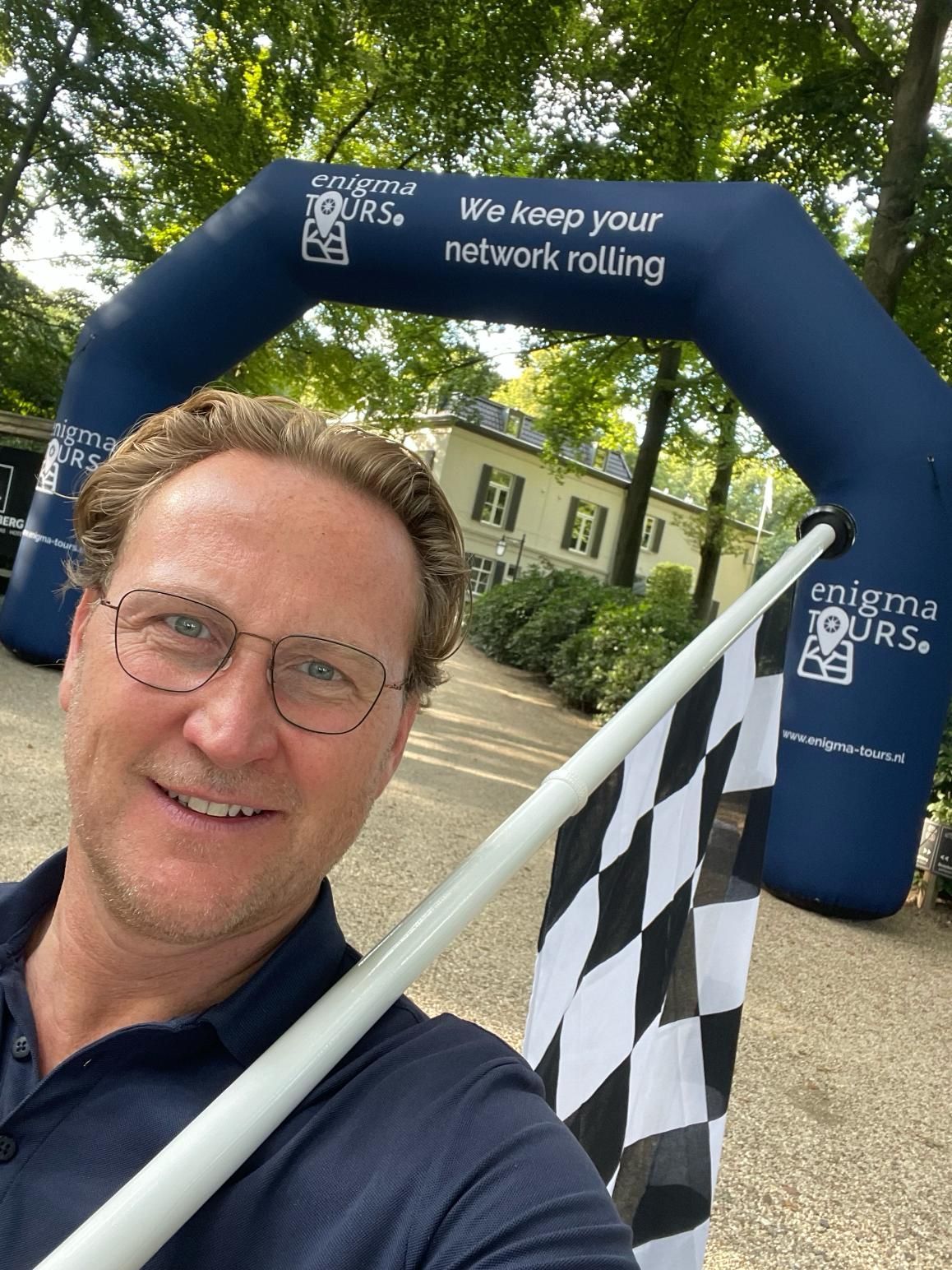 Man met bril maakt een selfie voor een blauwe boog met de tekst 