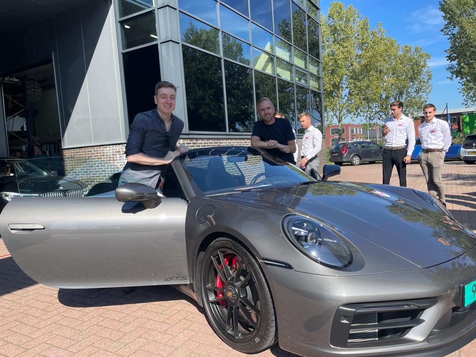 Drie mannen staan ​​naast een grijze Porsche cabriolet voor een gebouw. ​​Een van hen zit achter het stuur.