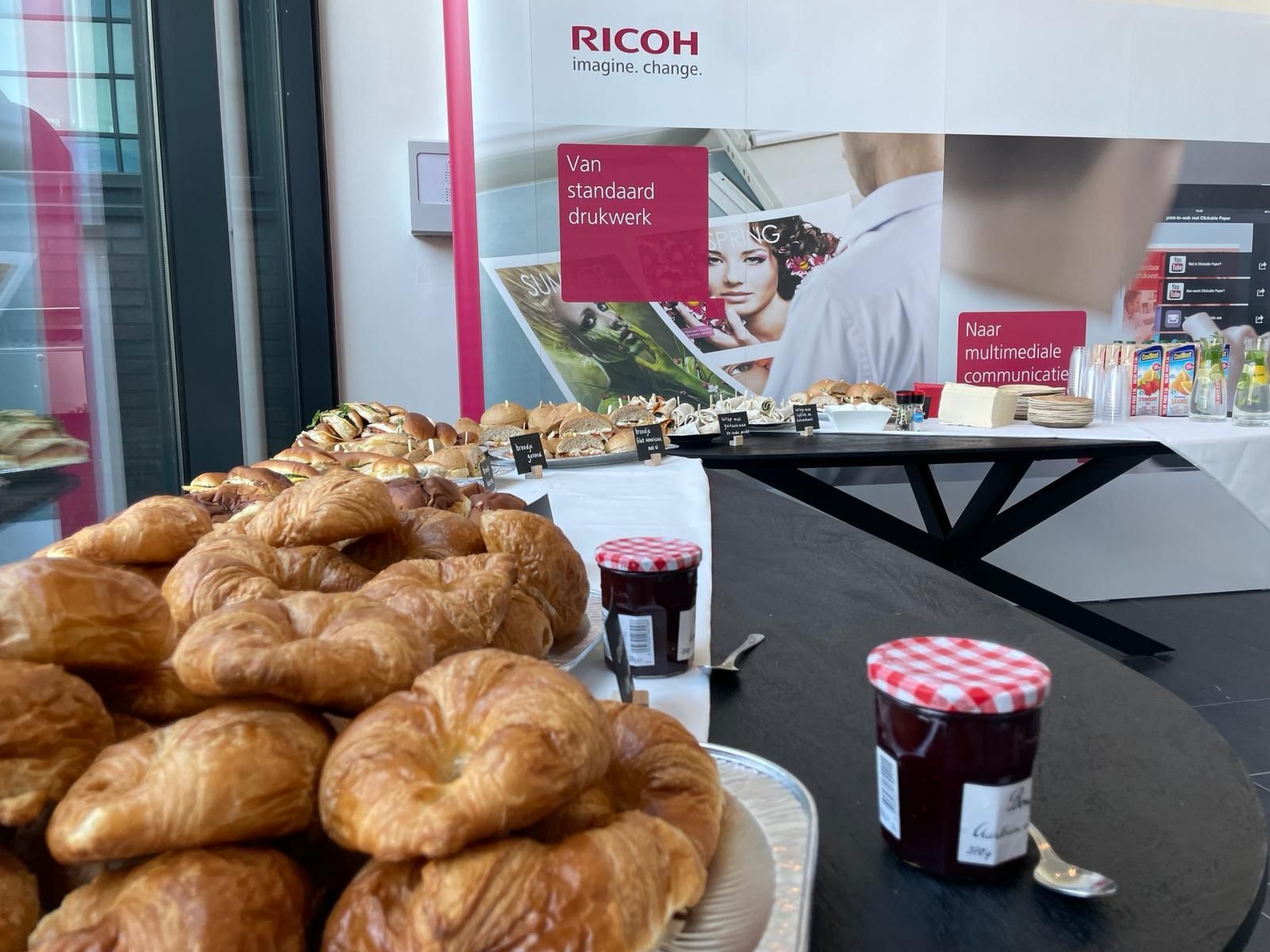 Een tafel met croissants, jampotten en ander eten, voor een Ricoh-banner.