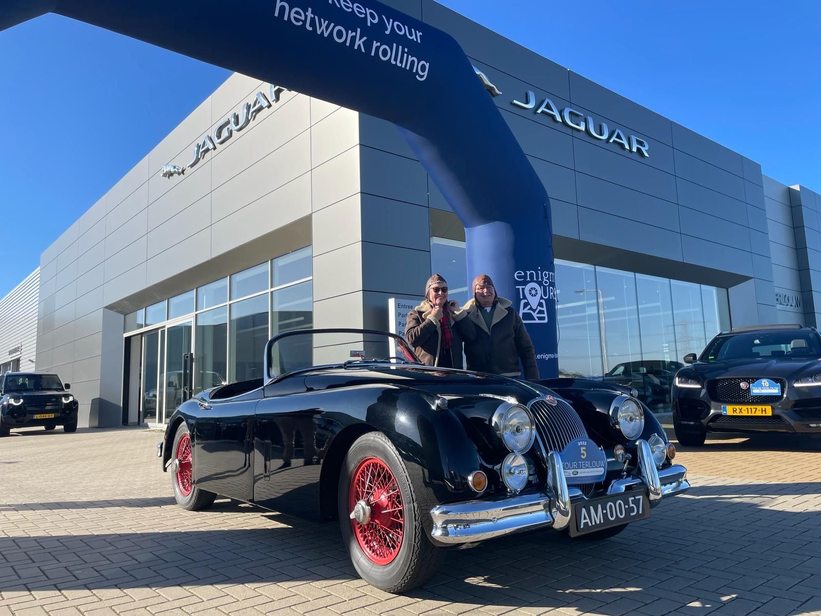 Twee mensen poseren naast een zwarte klassieke Jaguar cabriolet voor een Jaguar-dealer.