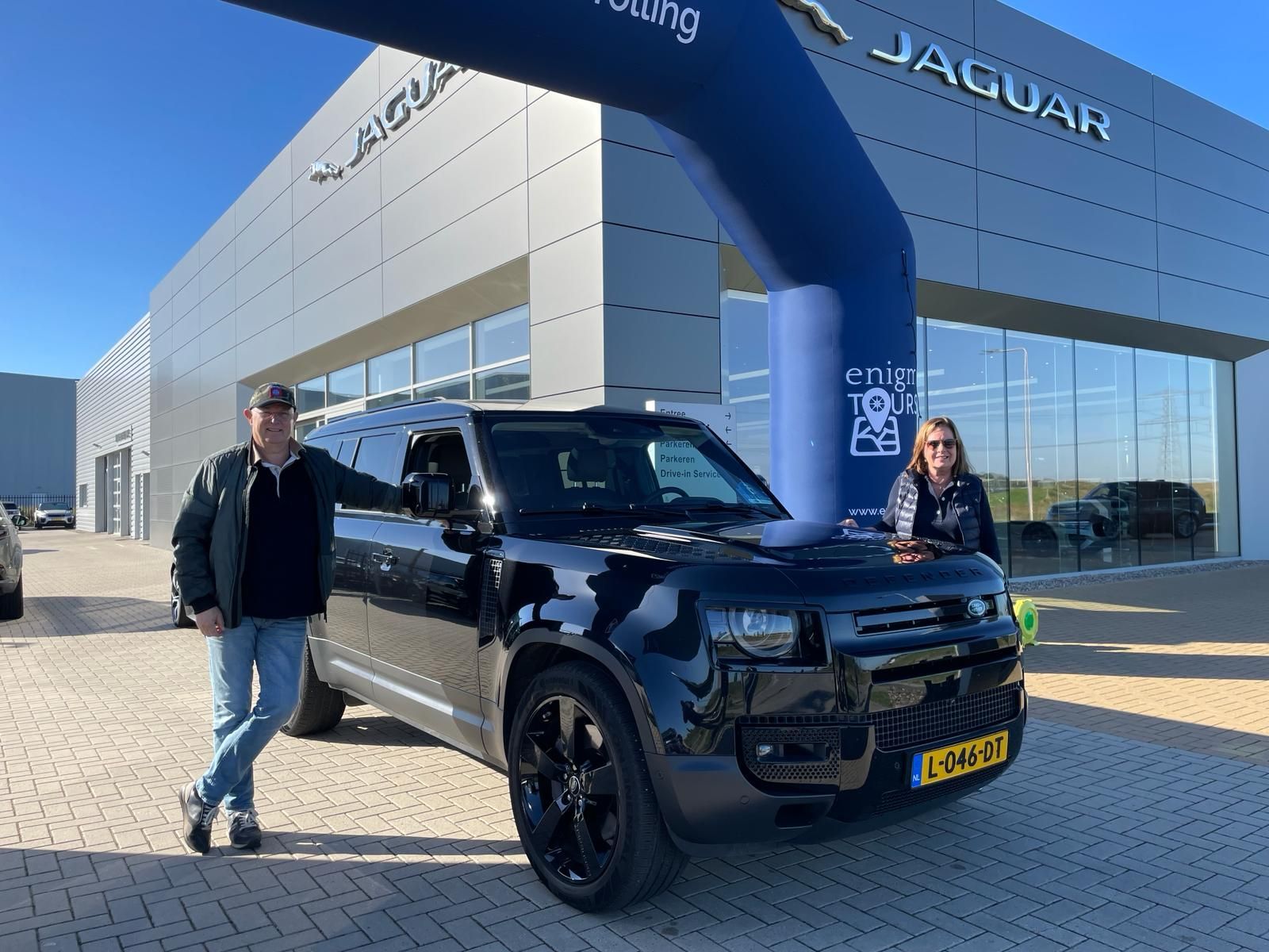 Twee mensen poseren met een zwarte Land Rover Defender voor een Jaguar-dealer.