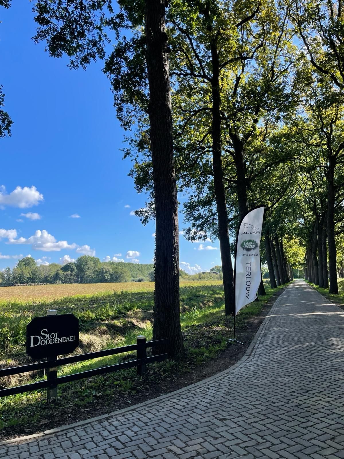 Een met bomen omzoomde bakstenen weg leidt naar een bord met de tekst 