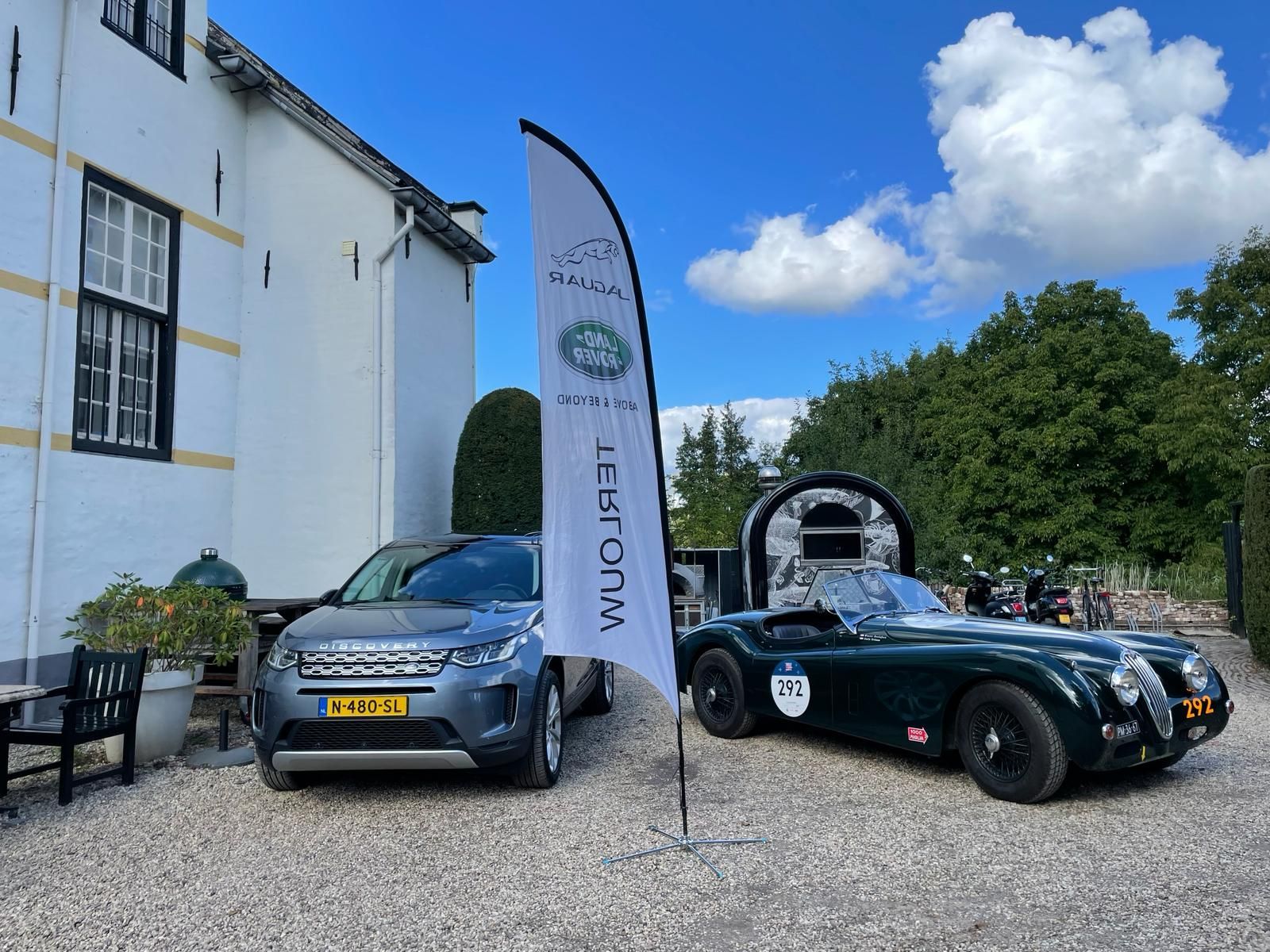 Twee auto's, een moderne SUV en een klassieke sportwagen, staan ​​geparkeerd bij een gebouw. ​​Tussen de twee auto's staat een vlag met een logo.
