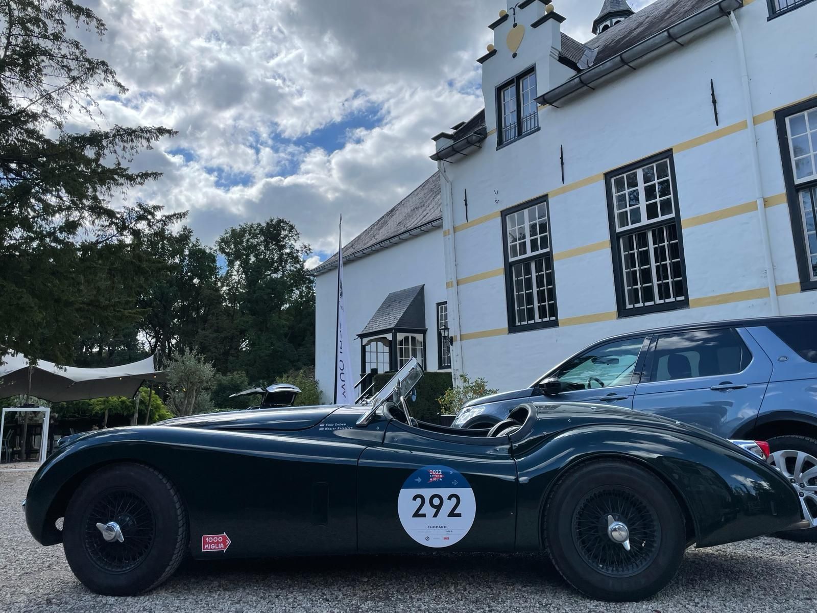 Een donkergroene vintage Jaguar sportwagen staat geparkeerd voor een wit gebouw met nummer 292 op de deur.