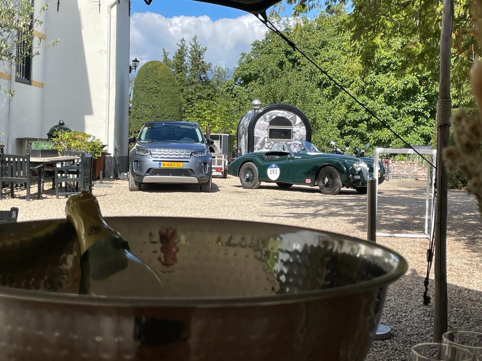 Zilveren emmer met champagne op de voorgrond; Land Rover en groene sportwagen op de binnenplaats.