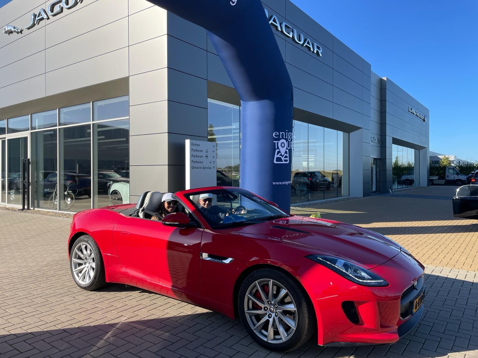 Een rode Jaguar cabriolet verlaat een autodealer met twee inzittenden.