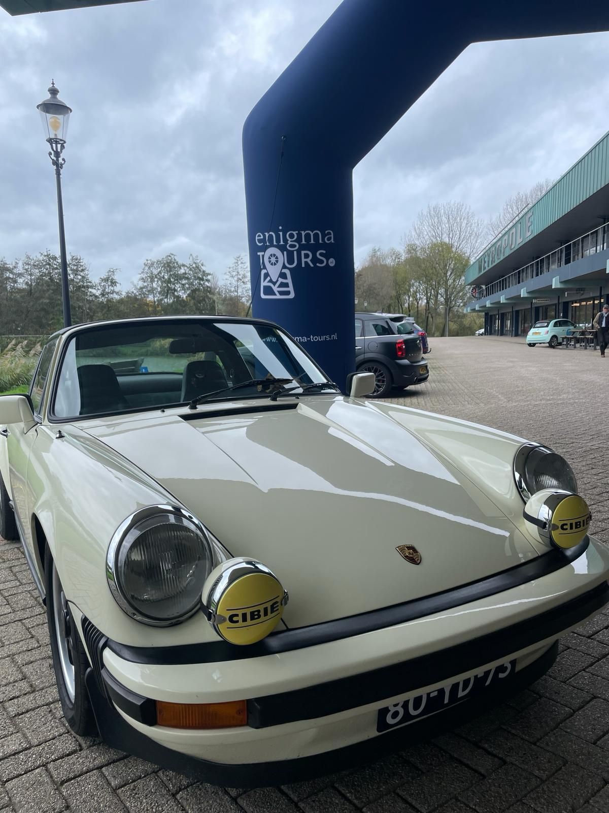 Crèmekleurige Porsche 911 met gele mistlampen, geparkeerd bij een blauwe poort, exterieurfoto.