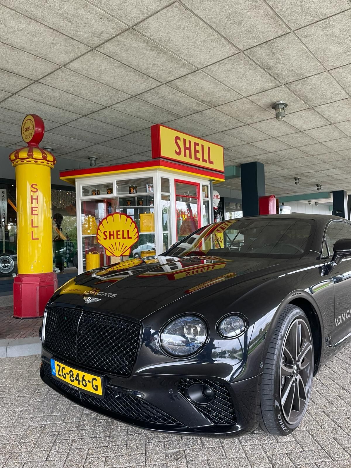 Een zwarte Bentley staat geparkeerd bij een Shell-tankstation met gele en rode borden.