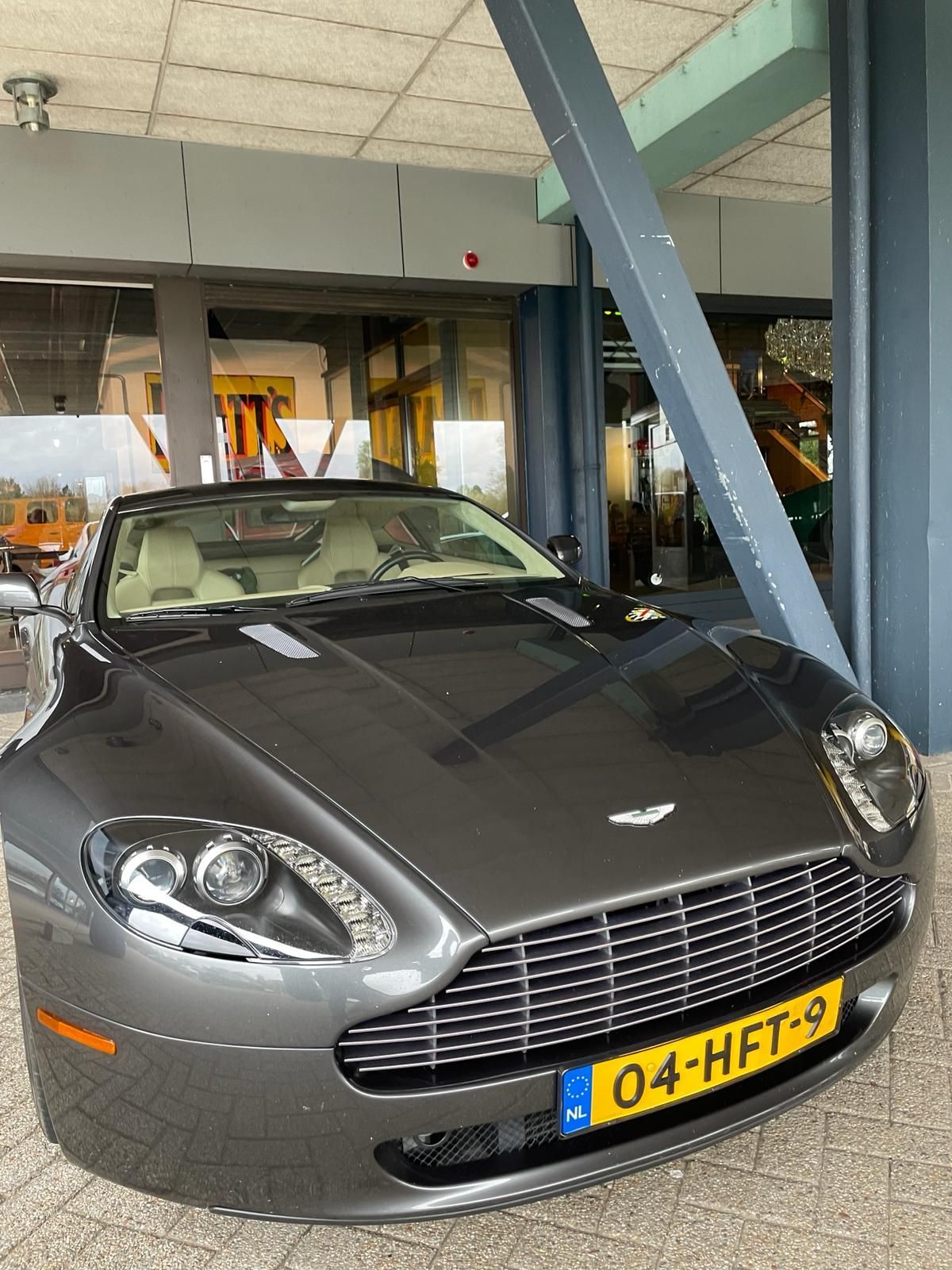 Grijze Aston Martin cabriolet geparkeerd onder een gebouw met Nederlands kenteken.