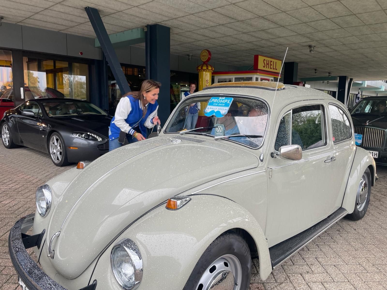 Een beige Volkswagen Kever staat geparkeerd op een autoshow, een vrouw kijkt ernaar, met andere auto's op de achtergrond.