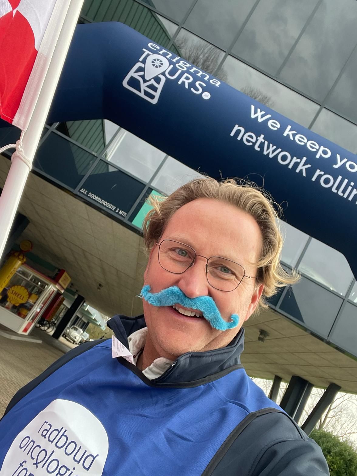 Man met blauwe snor, gekleed in een vest en met een bril, voor de boog van 