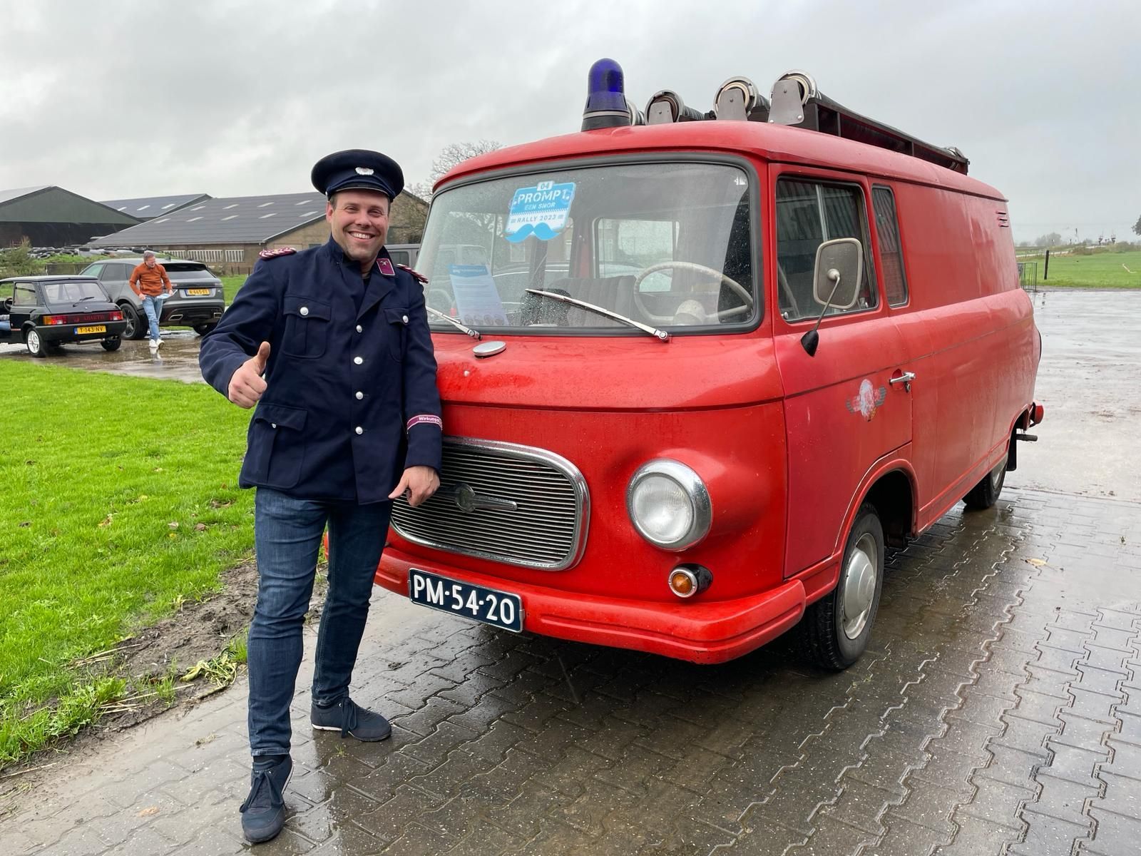 Een man in uniform steekt zijn duim omhoog naast een rode oldtimer brandweerwagen met blauw zwaailicht. Buiten, bewolkte dag.